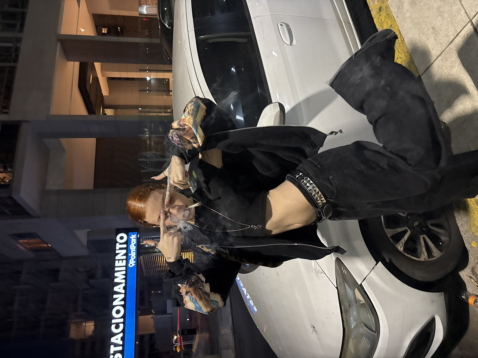 An individual with striking red hair is captured in an urban setting at night, leaning casually against a white car. The person, who appears female, is wearing a black cropped top, a black open hoodie with colorful graphic sleeves, black wide-leg pants, and large black boots. A prominent studded belt adorns their waist, and a long silver chain necklace hangs around their neck. Tattoos are visible on their right wrist.
In the midst of smoking a cigarette, with plumes of smoke drifting from their mouth, the person looks directly at the camera. Their right hand is raised, making a two-finger gesture near their eye, possibly a peace sign or a stylized pose. The background features a modern building with illuminated windows, suggesting an active cityscape.
On the left side of the frame, a tall vertical sign is clearly visible. It displays the Spanish word "ESTACIONAMIENTO" (meaning "PARKING") in white capital letters on a black background. Below this, a blue section of the sign features "PointPark" with an associated logo. The ground around the car shows yellow markings, characteristic of a parking area or roadside. The scene is set in Buenos Aires, Argentina. The overall mood is cool and casual, with the person exuding an edgy aesthetic.