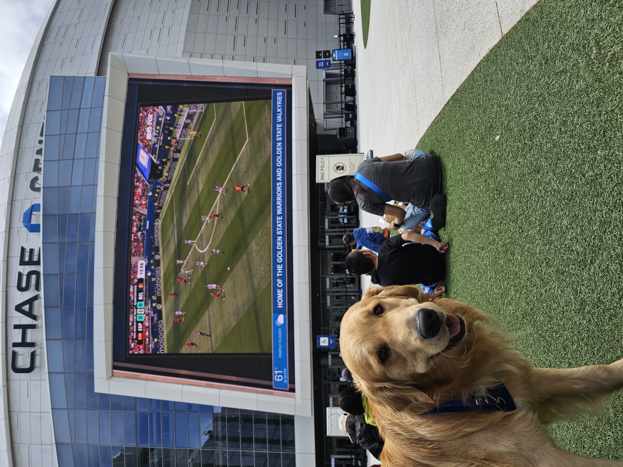 A golden retriever dog with a blue bandana stands in the foreground on artificial turf. In the background, a large LED screen displays a soccer match, with the score "ESP 0 - 0 BEL" and the time "13:56". The screen also shows "FIFA WORLD CUP 2026™ QUARTERFINAL" and "motorola" logos. Below the screen, text reads "61° PARTLY CLOUDY" and "HOME OF THE GOLDEN STATE WARRIORS AND GOLDEN STATE VALKYRIES". The stadium, identified as Chase Center, is visible behind the screen. Several people are sitting on the turf in the background, facing away from the camera. A "BAG POLICY" sign is also visible. The overall environment appears to be an outdoor plaza area of a sports arena in San Francisco, California, United States of America.