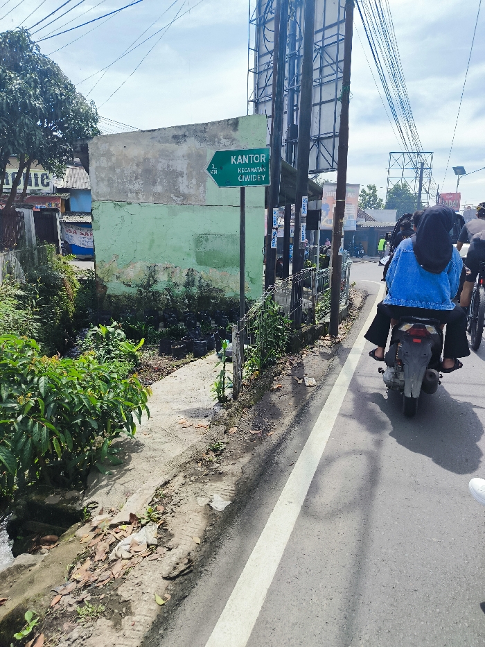 The image shows a street scene in Ciwidey, Indonesia, with a green sign that reads "KANTOR KECAMATAN CIWIDEY," which translates to "District Office Ciwidey." The sign is located on the side of the road, near a green building with a concrete walkway in front of it. There is a small, overgrown patch of green plants along the side of the walkway. A person wearing a blue jacket and a black hijab rides a motorcycle down the road. The road is paved and has a white dividing line. The road appears to be a single lane. The sky is clear and blue with some white clouds. The location of the image, based on the coordinates, is a residential area with a mix of houses and small businesses.