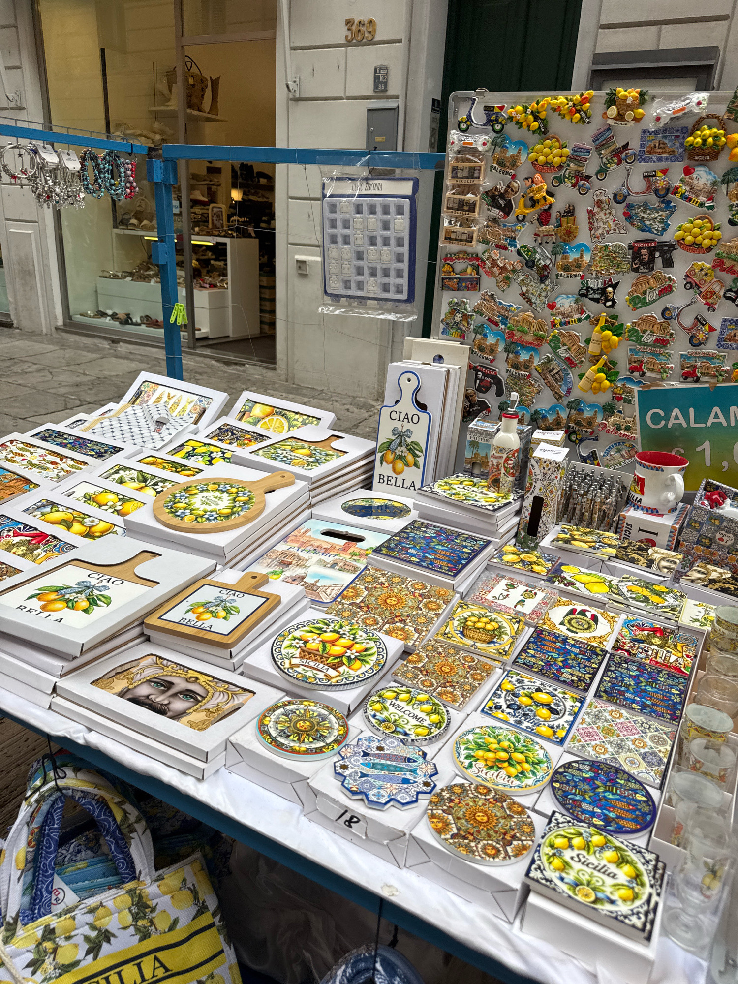 The image depicts a street market stall in Palermo, Italy, displaying a vibrant array of souvenirs. The stall is laden with items, primarily featuring ceramic coasters and decorative tiles adorned with traditional Sicilian motifs, prominently showcasing lemons and intricate patterns. Several small wooden cutting boards with similar designs are also visible. To the left, a blue metal frame supports a display of beaded bracelets. Behind the stall, a shop window displays clothing and accessories, suggesting a commercial area. The background reveals the building's facade with a street number "369" visible above the shop entrance. The overall scene is a bustling outdoor market, indicative of a tourist-frequented area in Palermo.