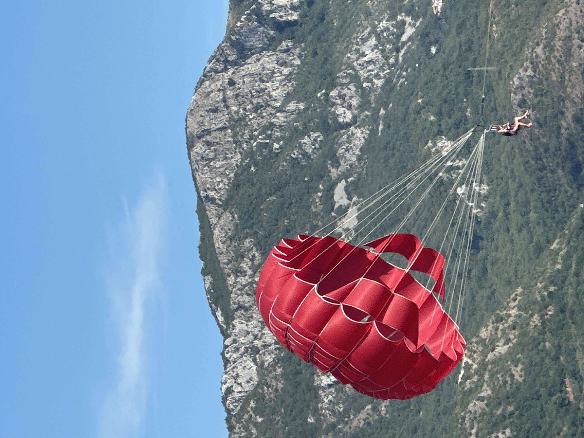 A red parachute with a person parasailing is in the air, with a mountainous landscape in the background. The parachute is inflated and dominant in the foreground, with its lines extending down to the parasailer. The parasailer is seated in a harness and appears to be enjoying the activity. The background consists of a steep, rocky mountain covered with green vegetation. The sky is clear and blue. The location is Boreti, Montenegro.