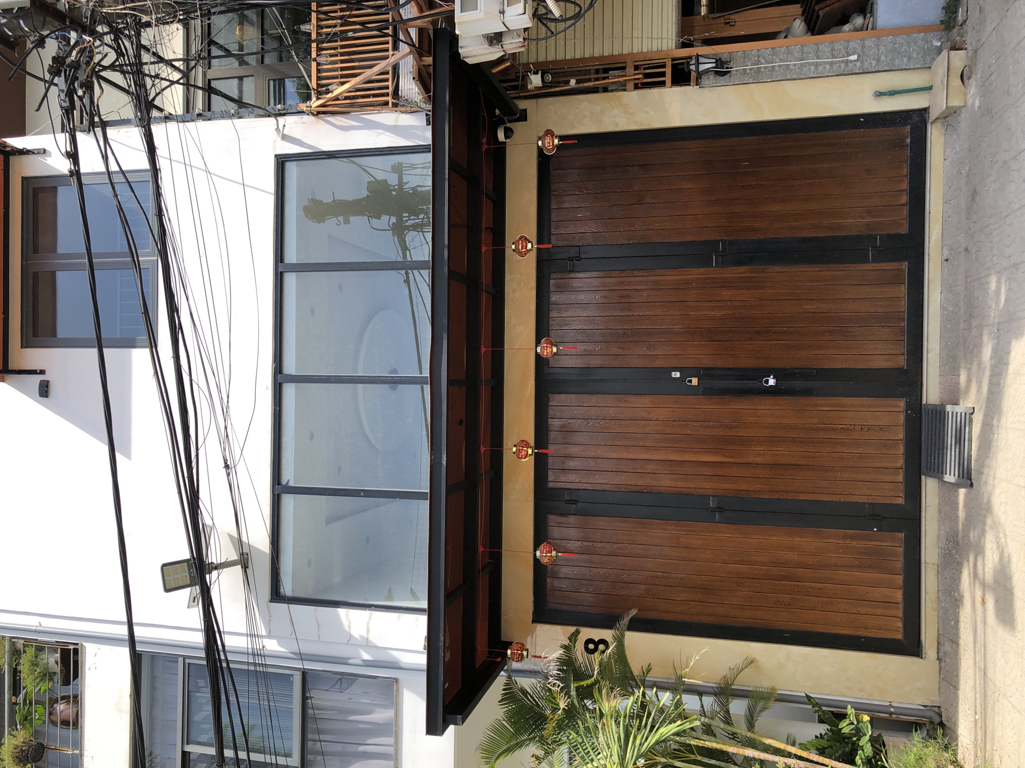 The image captures a building facade in Đà Nẵng, Vietnam. The building is multi-story, painted white, with large windows. The ground level features a large set of double wooden doors framed in black. Above the doors, a black awning extends outwards. Red lanterns are suspended from the awning, adding a decorative element. A single digit "8" is visible on the wall to the left of the doors. To the left of the entrance, a palm plant is present. The scene is characterized by an abundance of electrical wires running across the facade, suggesting an urban setting. Security cameras are also visible mounted under the awning.