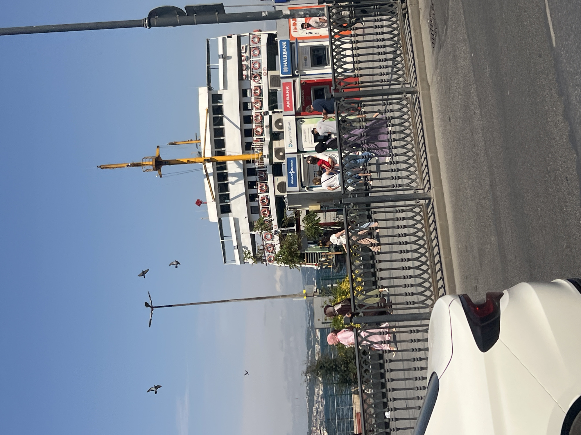 In Istanbul, Turkey, a white sedan is parked on the right side of the frame, facing left. The car's rear end is visible, with its taillight illuminated. Across a dark-railed promenade, people walk alongside a ferry. The ferry, a white, multi-deck vessel, is docked, and its mast is visible. Several ATM machines are situated near the ferry, with the logos of "Türkiye İş Bankası," "Garanti BBVA," "AKBANK," and "HALKBANK" displayed on them. The sky is clear blue, and birds are seen flying in the distance. The scene suggests a busy waterfront area, likely a ferry terminal or port in Istanbul.