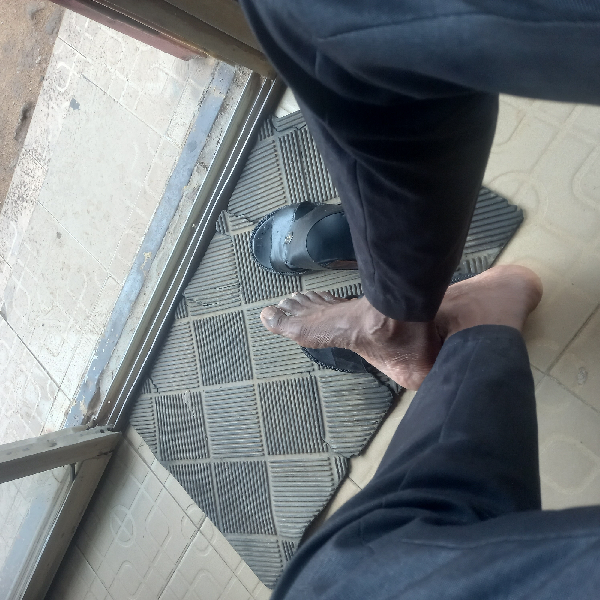 A top-down view depicts a person standing on a rectangular grey doormat. The person wears black trousers. The left foot is clad in a black sandal, while the right foot is bare. The doormat is positioned just inside a doorway, with a metal frame visible on the left. The floor inside is tiled in a light, patterned design. The setting appears to be an indoor entrance. The location is specified as Dadi Riba, Nigeria.