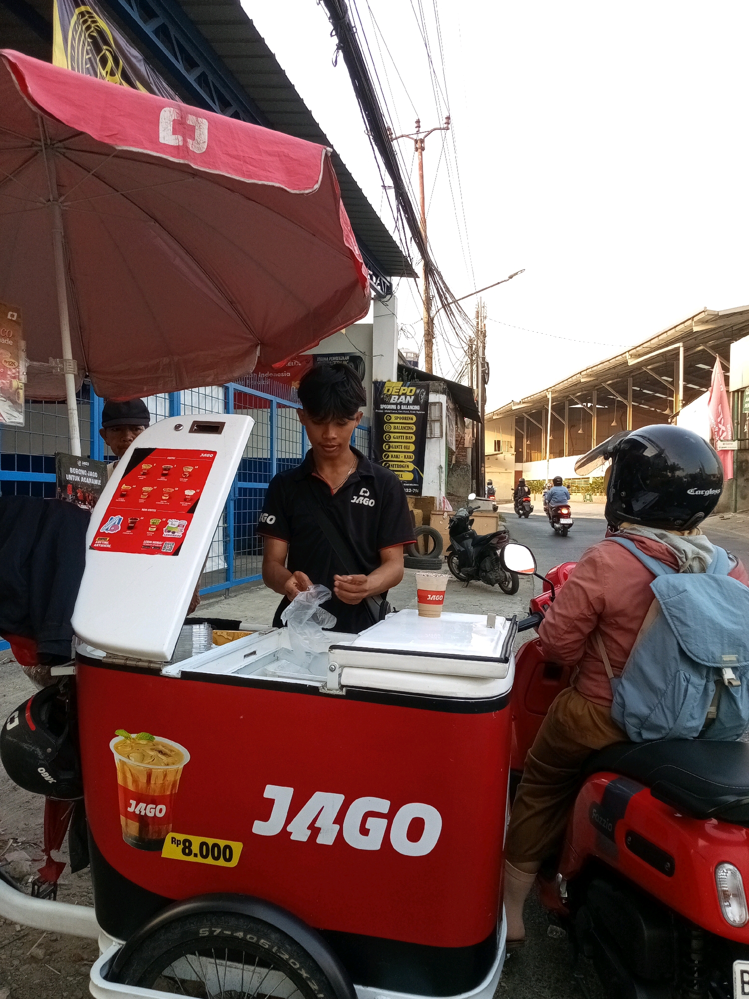 In the city of Bekasi, Indonesia, a street vendor is preparing a drink at a mobile cart with a red and white cooler. The cart is adorned with the logo "JAGO" and a picture of a beverage with the price "Rp 8.000". A young man, likely the vendor, is seen handling plastic packaging. Behind him, another man is visible. A customer on a red scooter, wearing a black helmet and a blue backpack, is waiting to purchase a drink. The scene is set against a backdrop of buildings and overhead electrical wires, suggesting an urban environment. The lighting indicates it is daytime, likely late afternoon given the warm tones.