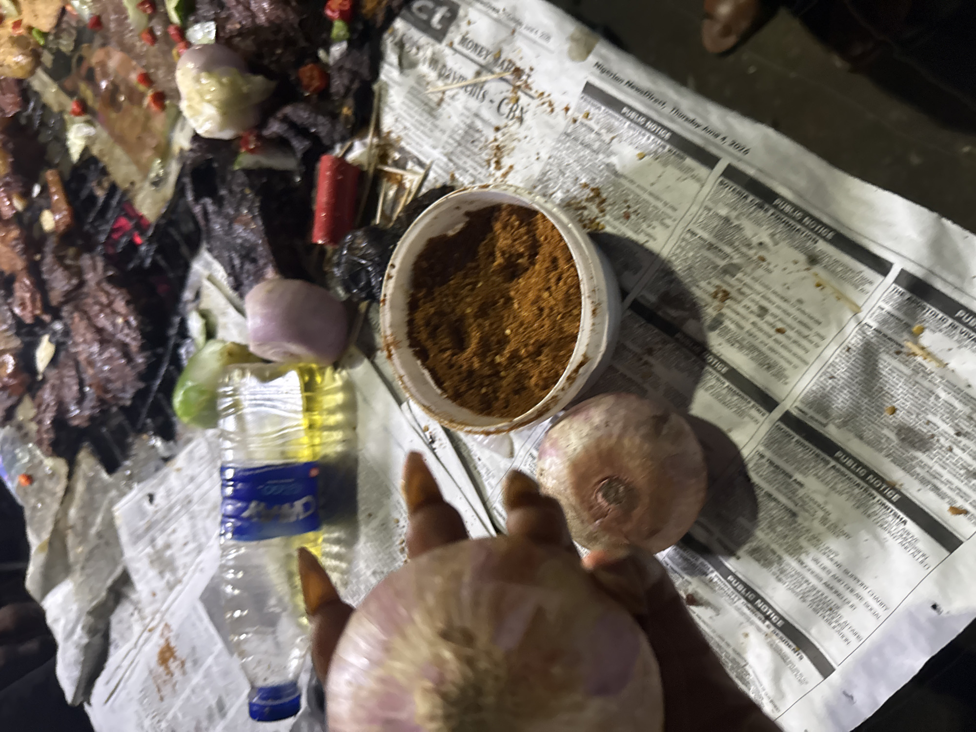 The image depicts a close-up of a person's hand holding two onions, with a bowl of ground spices in the foreground. In the background, there is a grill with cooked meat, a plastic bottle containing yellow liquid, and several chopped red chilies and onions. The items are placed on a surface covered with newspapers, some of which appear to be from the "Abuja Newspaper," suggesting the location may be Abuja, Nigeria. The overall scene appears to be an outdoor food preparation or serving area.