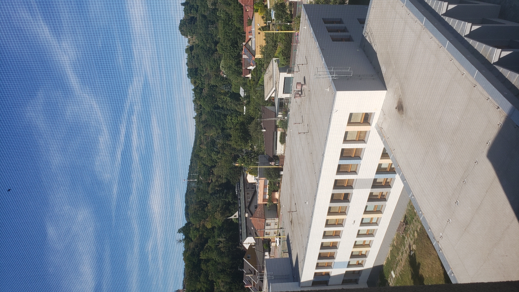 The image shows a view from a window of a building, looking out over a residential area with houses and trees, set against a backdrop of a green, tree-covered hill. The sky is bright blue with scattered white clouds. In the foreground, the flat, grey roof of another building is visible, with several vent pipes and some dark stains on its surface.
The main building in the mid-ground is a light-colored, multi-story structure with a grid of windows featuring light brown frames. Some of the windows have blue accents. The building appears to be a school or institutional building. Behind this building, a cluster of houses with red-tiled roofs and other buildings are visible, some with solar panels. The houses are surrounded by lush green trees and vegetation. The hill in the background is densely wooded, and a tall, slender white structure, possibly a communication tower, is visible near the top of the hill.
There are no people visible in the image. The overall scene depicts a daytime view of a semi-urban or suburban environment.
This scene is identifiable as the area near the primary school of ZŠ Šrobárova in Prešov, Slovakia. The specific building in the foreground is likely the ZŠ Šrobárova itself, viewed from another part of the school complex or a neighboring building. The arrangement of houses and the forested hill in the background are characteristic of the landscape in that vicinity of Prešov, which is situated at the edge of the city and bordered by hills.