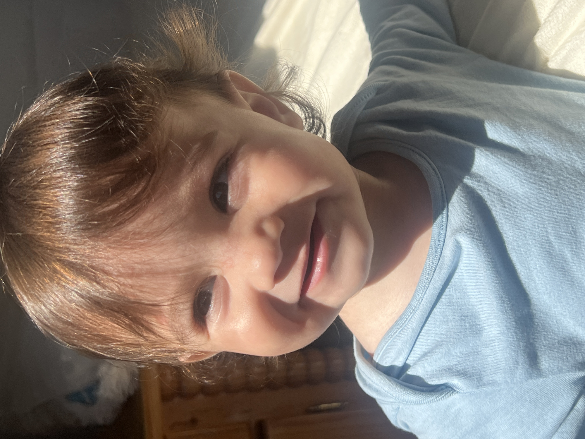 A young child with light brown hair and brown eyes is lying down, smiling. The child is wearing a light blue shirt. The background is blurry but appears to include a wooden surface. The scene is brightly lit with natural light. No information about location in Elena, Bulgaria is discernible from the image.