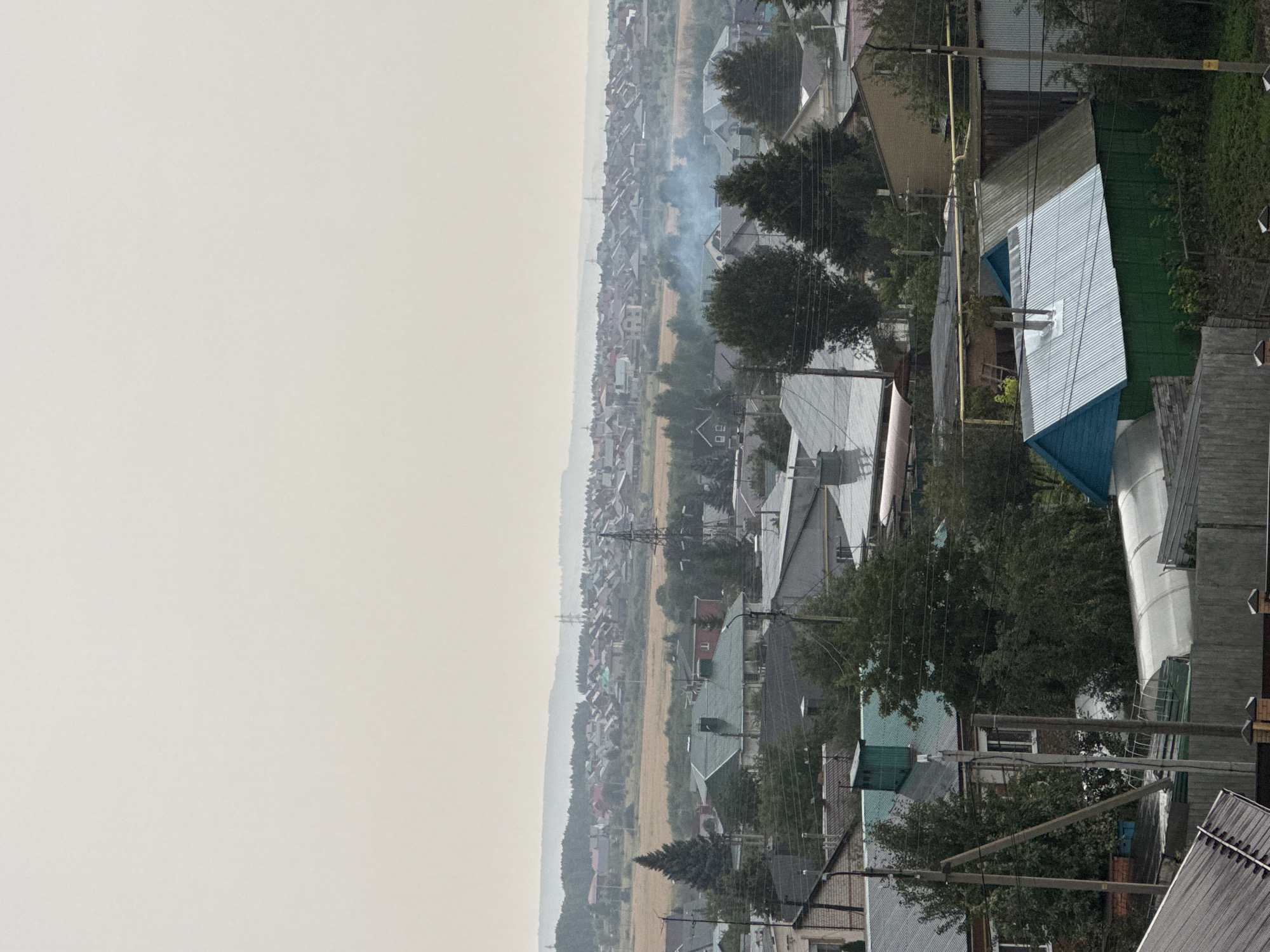 The image shows a hazy, overcast day in a suburban area of Almetyevsk, Russia. The foreground is dominated by the roofs of several residential buildings, varying in material and color, including corrugated metal and shingles. Some buildings have visible chimneys and vents. A greenhouse structure is also present in the foreground. Power lines and poles are a prominent feature, crisscrossing the view.
In the mid-ground, more houses are visible, interspersed with trees. A field of dry grass or crops stretches across the middle distance, behind which a dense line of trees forms a natural boundary. Further in the background, a vast expanse of similar residential rooftops extends towards the horizon, punctuated by occasional power pylons. A thin plume of smoke is visible rising from one of the houses in the mid-ground, indicating some ongoing activity. The sky is a uniform pale gray, suggesting diffused light conditions and possibly pollution or mist. The overall impression is a densely populated residential area undergoing typical daily life.