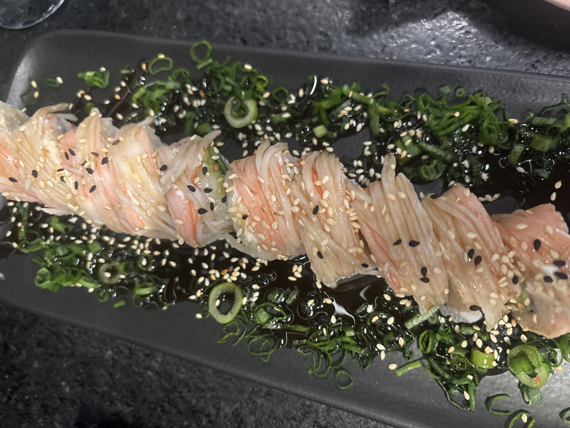 A close-up shot shows a rectangular black plate with a line of sushi rolls topped with shredded crab meat, sesame seeds, and black sesame seeds. The sushi is surrounded by chopped scallions and a dark, glossy sauce. The background is dark and out of focus, suggesting an indoor setting. The location is in San Bernardino Tlaxcalancingo, Mexico.