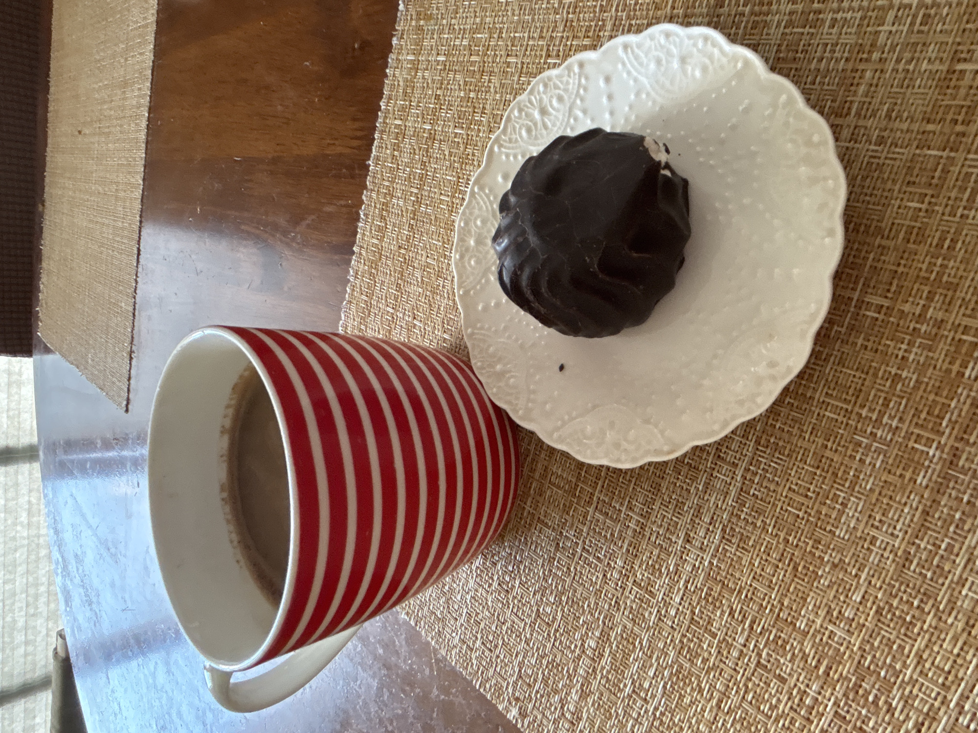 The image shows a close-up view of a red and white striped mug filled with a dark beverage, likely coffee or hot chocolate. Next to the mug, on a white decorative plate, sits a chocolate-covered marshmallow confection. Both the mug and the plate are placed on a woven straw placemat, which rests on a polished wooden table. The background is slightly blurred, but suggests an indoor setting with natural light filtering through blinds. There are no people visible in the image. The scene depicts a simple treat being enjoyed in a relaxed setting. The location in the city of Sochi, Russia, is not explicitly indicated by any visual cues within the image itself.