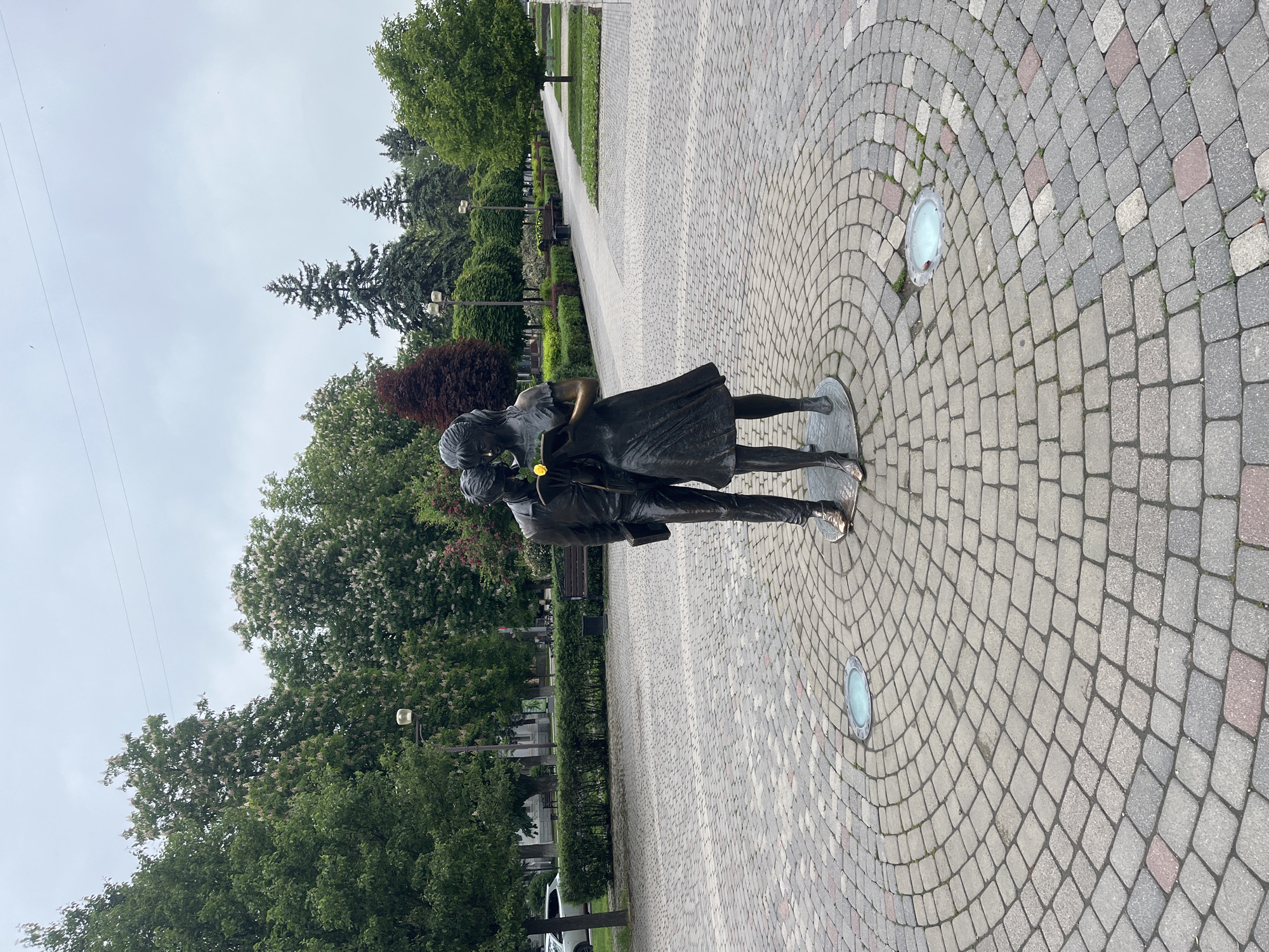 The image shows a bronze statue of a couple embracing in Krasnodar, Russia. The statue is positioned on a circular section of a cobblestone walkway with integrated round lights. The walkway is bordered by manicured hedges and trees. Beyond the hedges, a pedestrian path and benches are visible. The sky is overcast.