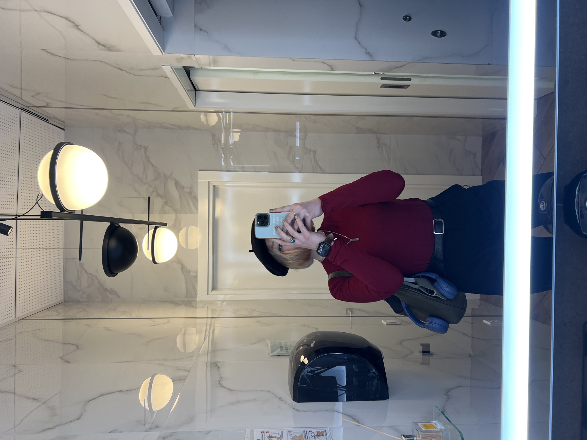 This media file captures a person taking a mirror selfie in a modern, well-lit indoor environment, identified as being in Odesa, Ukraine. The image is rotated 90 degrees clockwise.
The subject is a person, likely female, with short blonde hair and a black beret. They are wearing a red long-sleeved top and dark pants or a skirt with a black belt featuring a silver buckle. On their left wrist, a smartwatch is visible, and a ring adorns their right hand. They hold a light green iPhone with a dark case, positioned to take the photograph, partially obscuring their face. Blue over-ear headphones are draped over a bag, visible on their left side.
The setting features glossy white marble-patterned tiles covering both the walls and floor, contributing to a clean and reflective aesthetic. The space is brightly illuminated by various modern fixtures: a multi-bulb chandelier with spherical lights (some glowing white, one black), a prominent vertical light bar along the right edge of the mirror, and reflected recessed ceiling lights.
Other visible objects include a black automatic hand dryer mounted on the wall. A glass shelf beneath the hand dryer holds small bottles (possibly perfume or a diffuser) and small cards or papers, one displaying an illustration of hands, likely indicating instructions. Reflections of a white door or partition are also visible in the mirror.
The primary activity is a person engaged in taking a mirror selfie. The overall impression is that of a contemporary and stylish public or commercial restroom or changing area.
Visible text includes "DON'T TOUCH MY PHONE" (inverted) on the back of the person's phone case, and partially visible, indistinct, inverted text on the hand dryer.