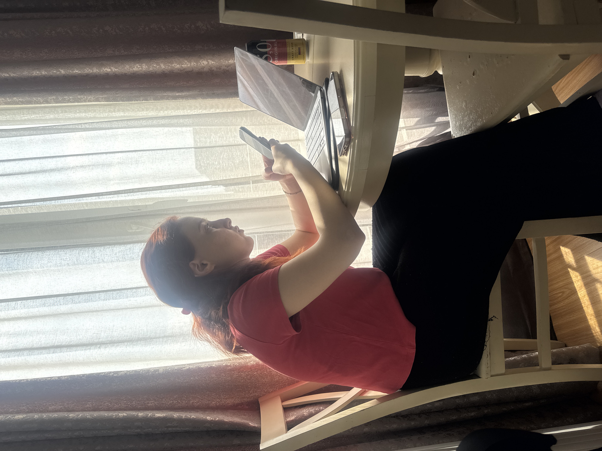A fair-skinned woman with reddish-brown hair sits at a white table. She is wearing a red t-shirt and black pants. The woman is looking at and holding a cell phone in her hands. A laptop and a can of beer are on the table. The backdrop features a window with blinds and a curtain. The location is potentially Tbilisi, Georgia, but there's insufficient information in the image to verify the city.