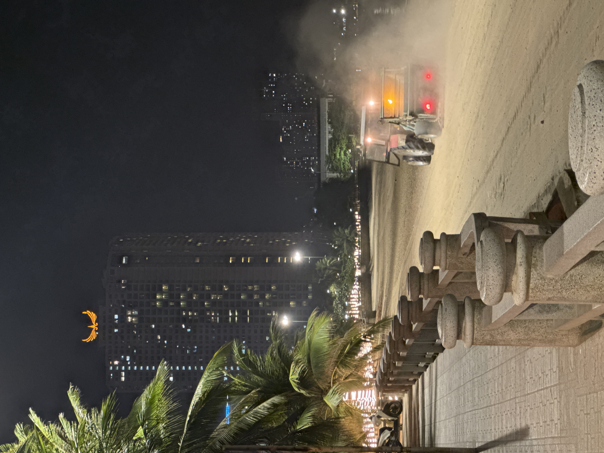 The image depicts a nighttime scene on a beach in Nha Trang, Vietnam. A compact tractor, equipped with a brush attachment and emitting dust, is actively cleaning the sandy surface. The tractor has its headlights and rear brake lights illuminated. In the background, a tall, modern high-rise building is visible, topped with a distinctive golden phoenix logo. Palm trees with illuminated strings of lights are present along the promenade to the left, adjacent to a paved walkway and a stone railing. The overall environment suggests an urban beach setting at night, with a focus on the maintenance of the beach area.