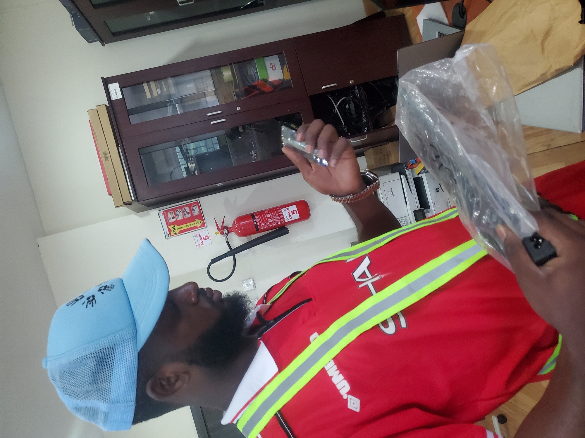 The image captures a dark-skinned man in an indoor setting, likely an office or storage room, in Gwagwada, Nigeria. He is wearing a light blue mesh trucker cap with a black patterned logo, a red short-sleeved polo shirt with a white collar and prominent neon yellow and grey reflective safety strips. A brown leather-strapped watch is visible on his right wrist. He has a full beard and appears focused on objects in his hands.
In his right hand, he holds a small, metallic, silver-colored object, possibly a component or tool. His left hand holds a larger item, obscured by clear plastic wrapping, suggesting it could be new equipment or a device.
The background features a dark wooden cabinet with glass doors, containing various items including boxes, cables, and what appears to be electronic gear, with one box inside clearly marked "5G". Stacked on top of the cabinet are several cardboard boxes. Mounted on the light-colored wall to the left of the cabinet is a red fire extinguisher labeled "FIRE EXTINGUISHER," "ADMIN 5," and instructions to "PRESS DOWN LEVER." A red and white safety sign is partially visible next to it. Further back, a white printer is discernable on a surface. The man's red shirt also shows a partial "UMBRO" logo and some indistinct text.
The scene depicts a functional environment, with the man seemingly engaged in an activity related to handling, inspecting, or setting up equipment. The image is rotated 90 degrees clockwise from its original orientation.