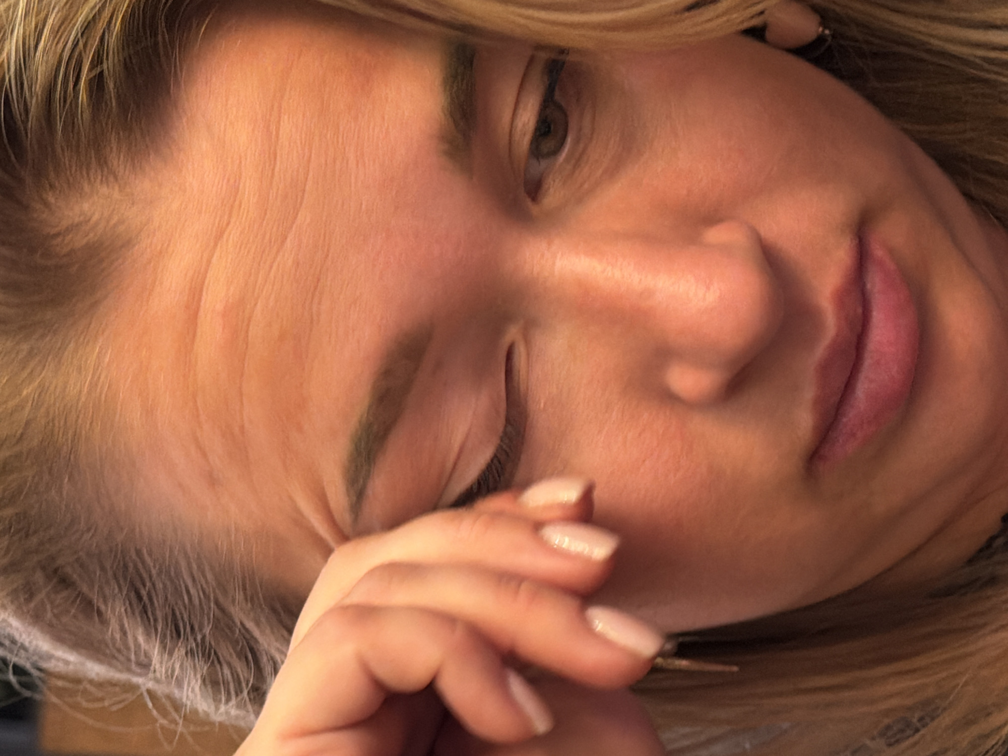 The image is a close-up, medium shot of a woman's face. She has blonde hair and is looking down and to the left. Her right hand is raised to her face, with her fingers touching her right eye. She is wearing light-colored nail polish. There is a subtle blush on her cheeks and pink lipstick on her lips. She is wearing a small, gold earring in her right ear. The background is blurred, but appears to be indoors with warm lighting. There are no visible landmarks or contextual clues to suggest a location in Unknown, Russia.