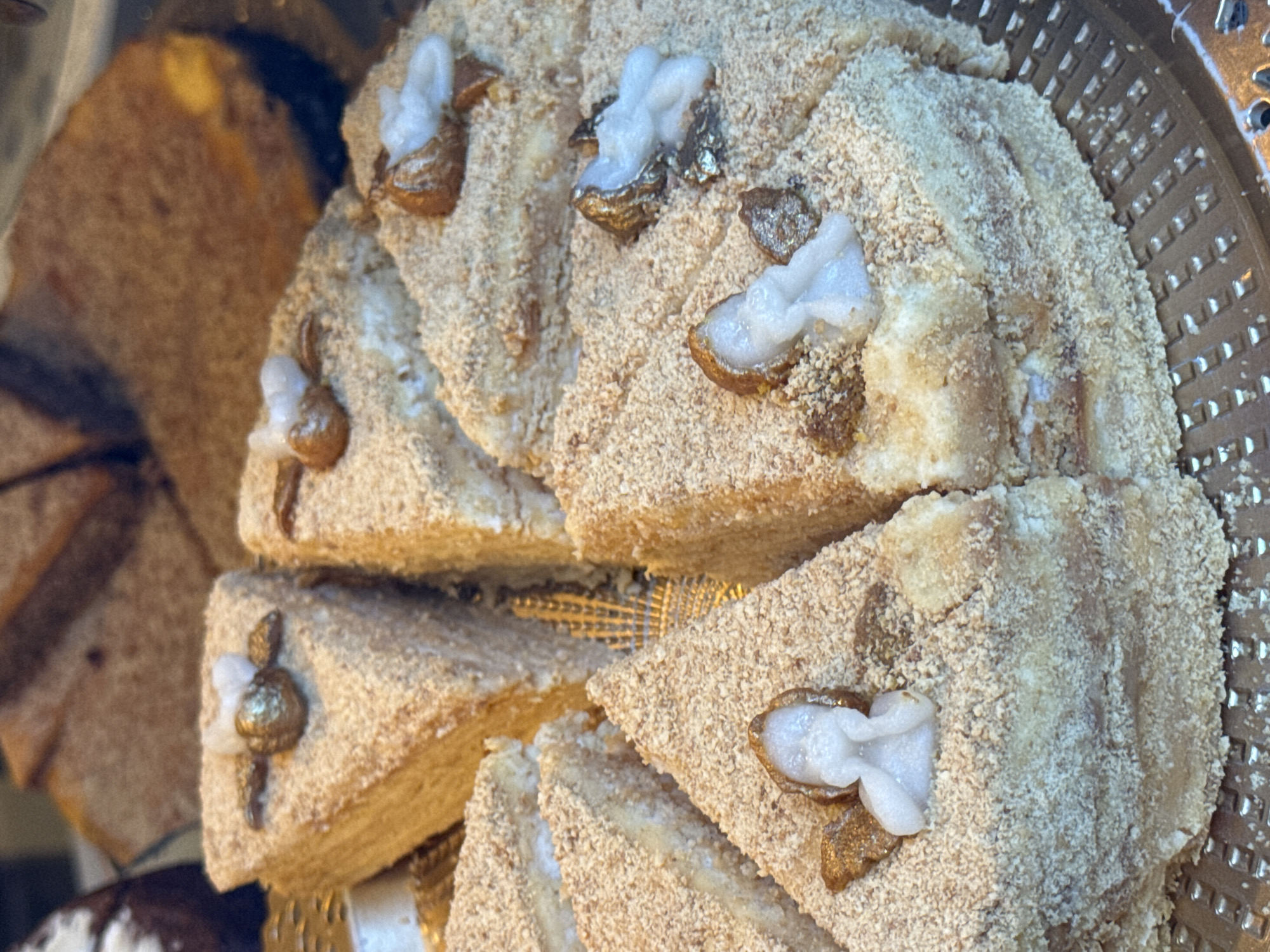 The image showcases a close-up view of a sliced cake, likely a Medovik (honey cake), displayed on a patterned golden tray. Each slice is topped with a decorative element resembling a white flower or figurine set in a shiny, metallic base. The cake's surface is covered with fine crumbs or ground nuts, giving it a textured appearance. The background includes additional slices of cake, possibly of different varieties. The scene appears to be set in a pastry shop or bakery, likely located in Barcelona, Spain, given the style and presentation of the desserts which are typical of European bakeries. The focus is on the visual appeal and intricate details of the cake, suggesting a focus on craftsmanship and culinary artistry. No people are visible in the frame.