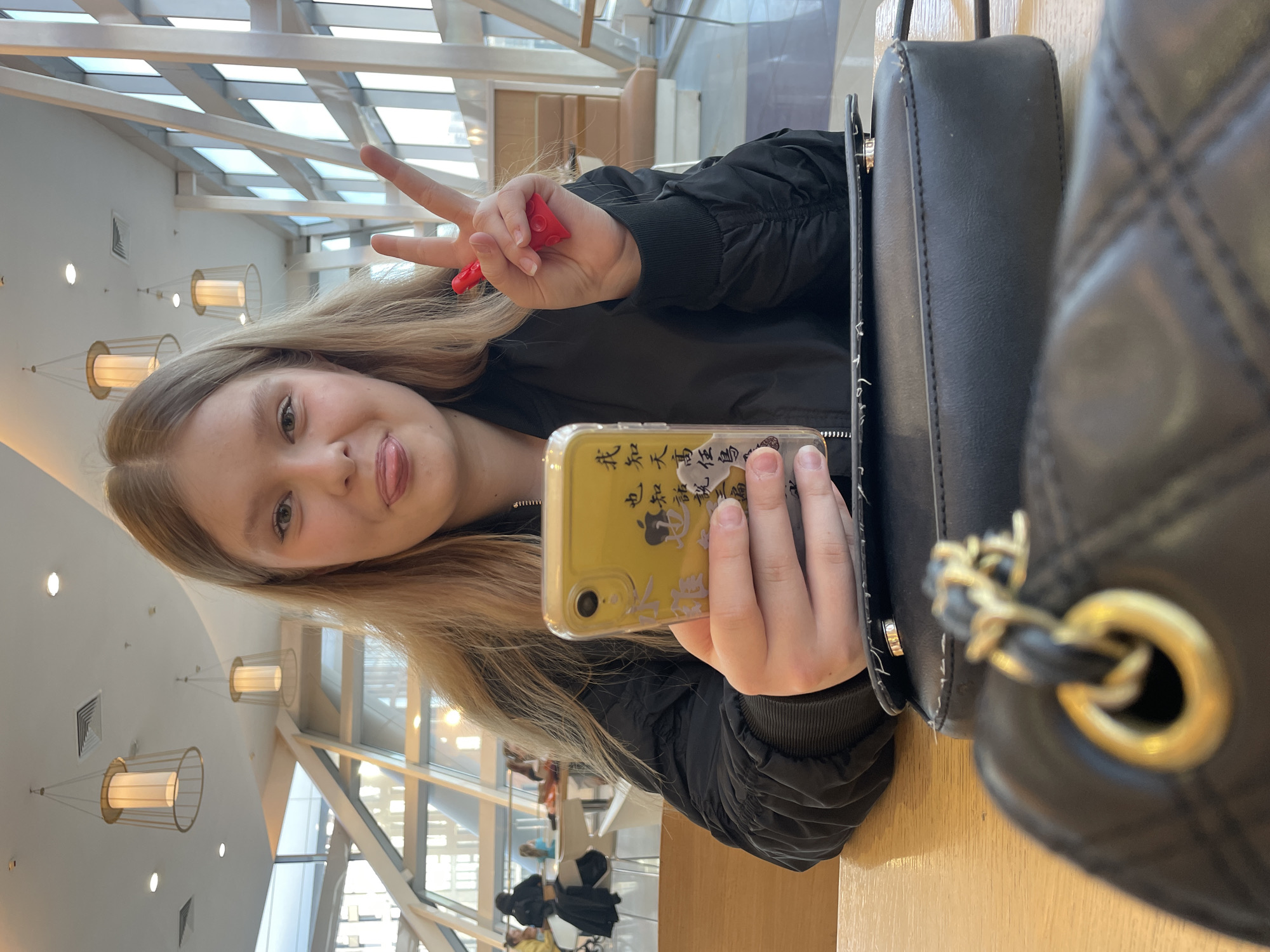 A young woman with blonde hair is taking a selfie indoors. She is holding a yellow-cased smartphone in her right hand and making a peace sign with her left hand, holding a red object. The background suggests a large indoor space with a high ceiling and many windows. Light fixtures hang from the ceiling. In the background, blurred figures suggest other people in the space. A black purse is placed on the table. The specific location cannot be determined.
