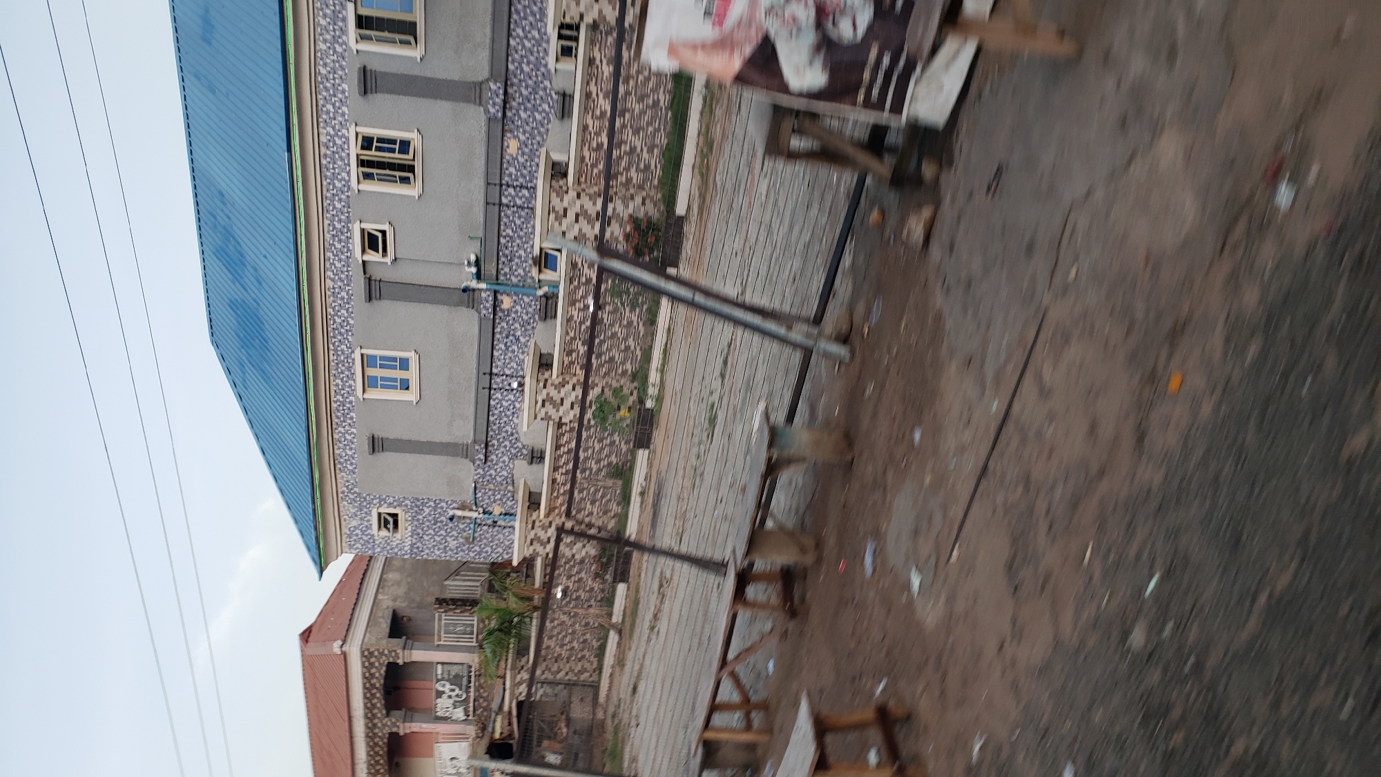 The image shows a street scene in Jalingo, Nigeria, with two buildings in the background. The building on the right has a blue, pitched roof and a grey facade decorated with a mosaic pattern. It has several windows, some open and some closed, with yellow frames. The building on the left has a reddish, pitched roof and a facade with columns and decorative elements. In the foreground, there are several wooden benches and a metal fence. The ground is dirt and appears to be littered with some debris. A large poster or banner is visible on the right side of the image, partially obscuring the fence and bench. The sky is overcast.