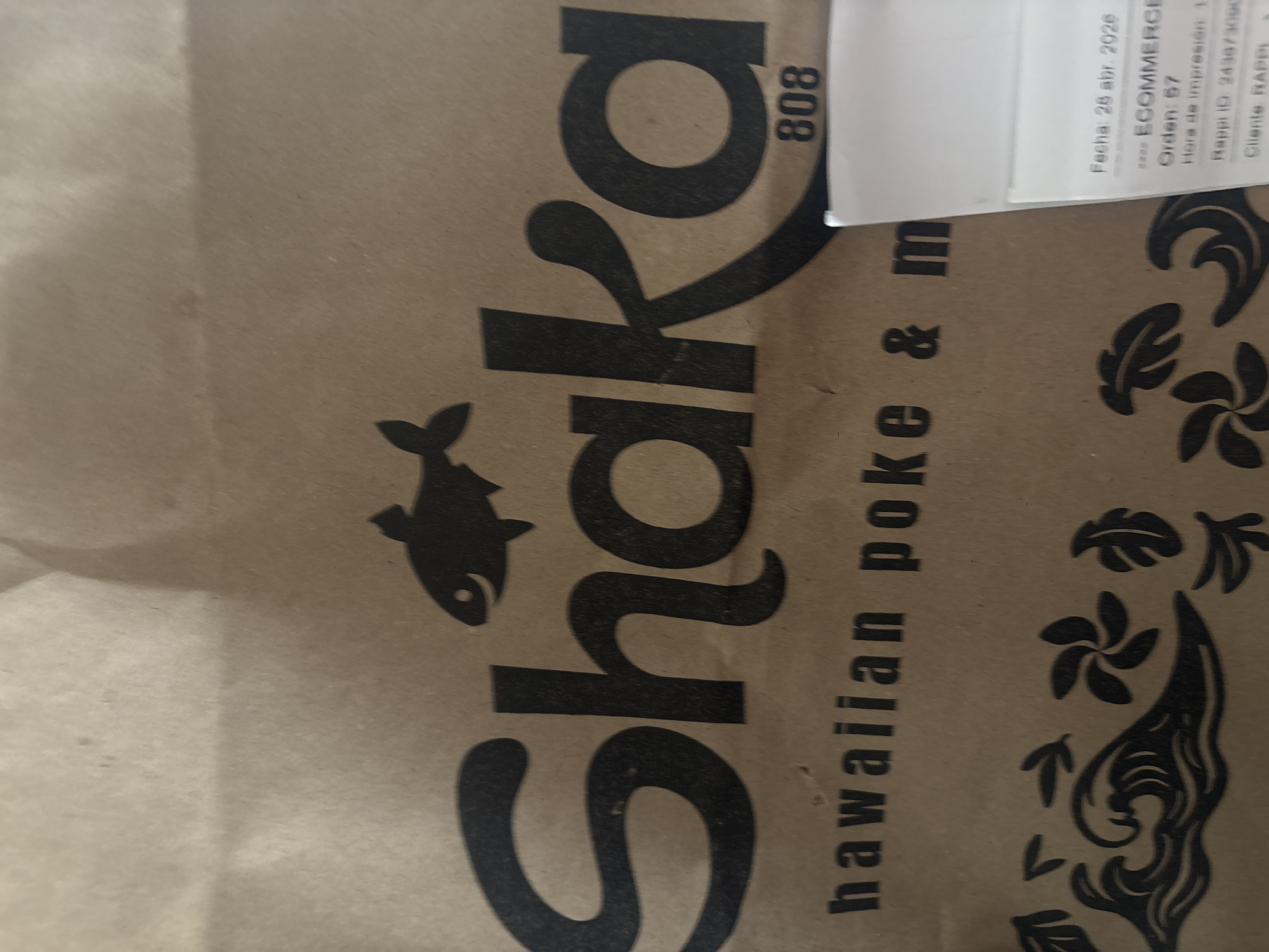 The image captures a close-up view of a brown paper bag, likely used for a takeaway or delivery order, positioned on an indistinct surface. The bag is prominently branded with the word "Shaka" in large, stylized black letters. To the left of the brand name, a black silhouette of a fish is visible. Below "Shaka," the text "hawaiian poke & m" is printed, suggesting "Hawaiian poke & more," indicating the type of food offered. Vertically placed next to the 'k' in 'Shaka' is the number "808." The right side of the bag is adorned with black illustrations of tropical leaves and flowers, further emphasizing the Hawaiian theme.
A white paper label, partially folded and obscured, is present in the upper right portion of the image. The text on this label provides details of an order, mostly in Spanish. Visible text includes "Fecha: 28 abr. 2026" (Date: April 28, 2026), "ECOMMERCE," "Orden: 57," "Hora de impresión: 1" (Print time: 1, partially obscured), "Rappi ID: 243073000," and "Cliente RAPP" (Customer RAPP, likely Rappi). These details indicate an e-commerce food order processed through the Rappi delivery service, a popular platform in Mexico City, Mexico. No people are present in the frame, and the setting appears to be indoors. The image itself is rotated, causing the text on the bag and label to appear sideways.