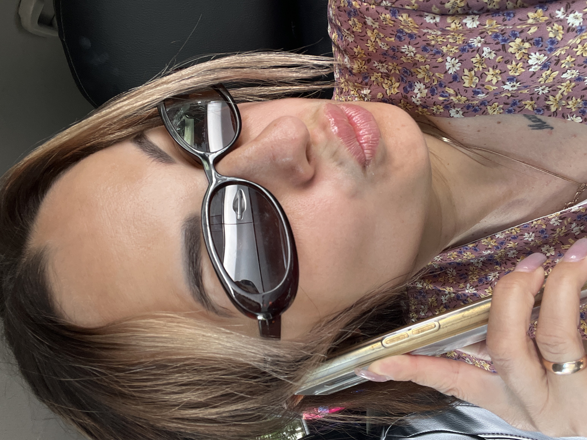 A woman is holding a golden smartphone to her ear, her lips pursed as if speaking or posing for a photo, while sitting in a vehicle. She is wearing sunglasses with dark lenses and a floral print dress. A ring is visible on her left hand. The background shows a dark car seat and a blurred reflection of a car in her sunglasses.