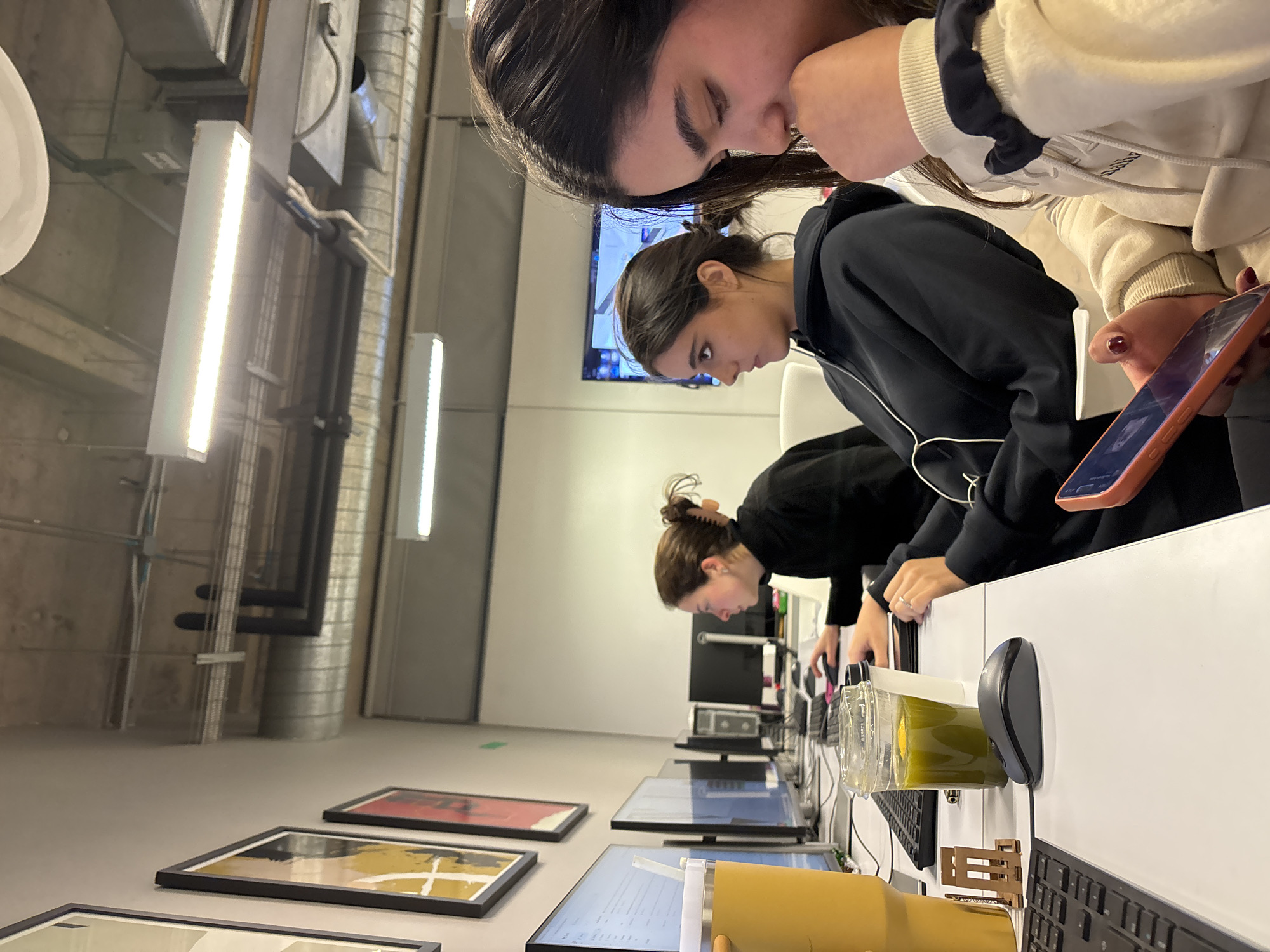 The image displays an interior view of an office or co-working space, likely located in Mexico City. Three young adult females are depicted at individual workstations.
In the immediate foreground, a female subject with long dark hair, wearing a light-colored hooded sweatshirt and a dark scrunchie on her right wrist, is observed looking downward, holding a pink-cased smartphone with her right hand. Her left hand is positioned near her chin.
To her left, in the mid-ground, a second female subject with dark hair pulled back, dressed in a black hooded sweatshirt and wearing white wired earbuds, faces forward and slightly right, away from the camera.
Further in the background, to the left, a third female subject with brown hair secured in a bun, wearing a black long-sleeved top, is seated at a computer workstation, actively using a computer mouse with her right hand.
The workstations feature white tabletops equipped with black computer monitors, keyboards, and mice. On the foreground desk, from left to right, a yellow insulated tumbler with a straw, a small wooden phone stand, a black wired computer mouse, and two beverage containers are visible: a clear plastic cup with a dome lid containing green liquid and a lemon slice, and a smaller, dark-colored cup behind it.
The environment includes light-colored walls, with three framed art prints (one yellow/orange, one red, one partially visible) adorning the left wall. The ceiling is exposed concrete, revealing metallic air ducts, utility conduits, and multiple rectangular LED strip light fixtures. A large screen or television monitor with indistinct content is visible in the far background.