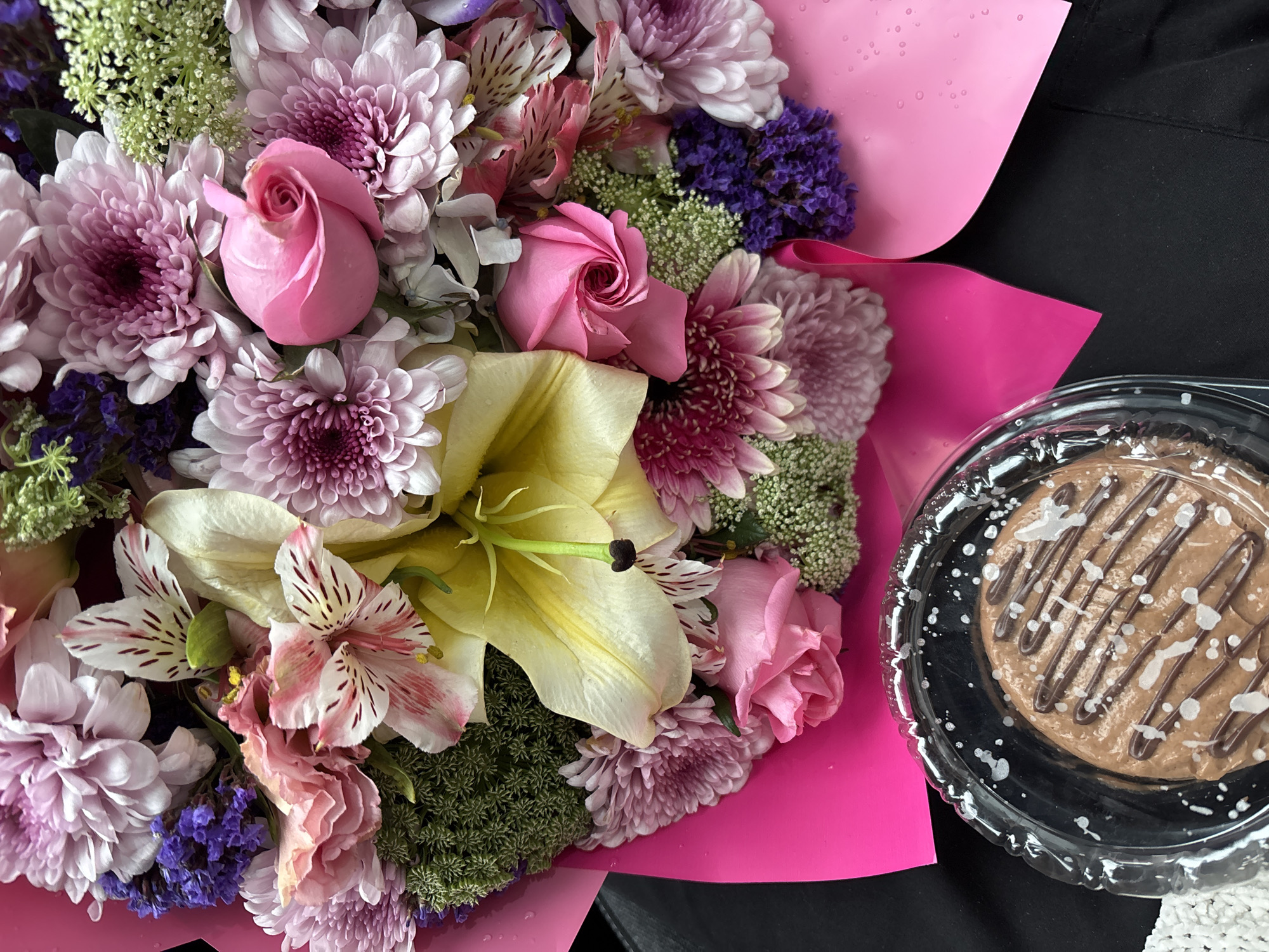 The image captures a vibrant still life arrangement featuring a large bouquet of fresh flowers and a small dessert. The bouquet, dominating the left side of the frame, is lavishly wrapped in bright pink paper, which shows numerous small water droplets, suggesting freshness.
The floral arrangement is rich and varied, showcasing an array of colors and textures. Prominent among the flowers are several pink roses, some still in bud, and numerous light purple and pink chrysanthemums with intricate petal structures. A large, open yellow lily stands out near the center, its vibrant color contrasting with the surrounding blooms. Other flowers include pink and white alstroemeria with distinct striped petals, clusters of delicate light green filler flowers, and sprays of deep purple blossoms, possibly statice.
To the right, positioned on a dark, possibly black, surface, is a single serving dessert enclosed in a clear plastic container with a fluted rim. The round dessert appears to be chocolate-flavored, with a brown base adorned with darker chocolate drizzle and scattered white speckles, possibly white chocolate or icing. A small section of white, textured fabric is visible in the bottom right corner, partially beneath the dessert container.
The scene, set in Puebla, Mexico, suggests a thoughtful gift or an indulgence for a special occasion. The lighting is soft and even, highlighting the details of the flowers and the dessert. There are no people visible, nor any discernible text.