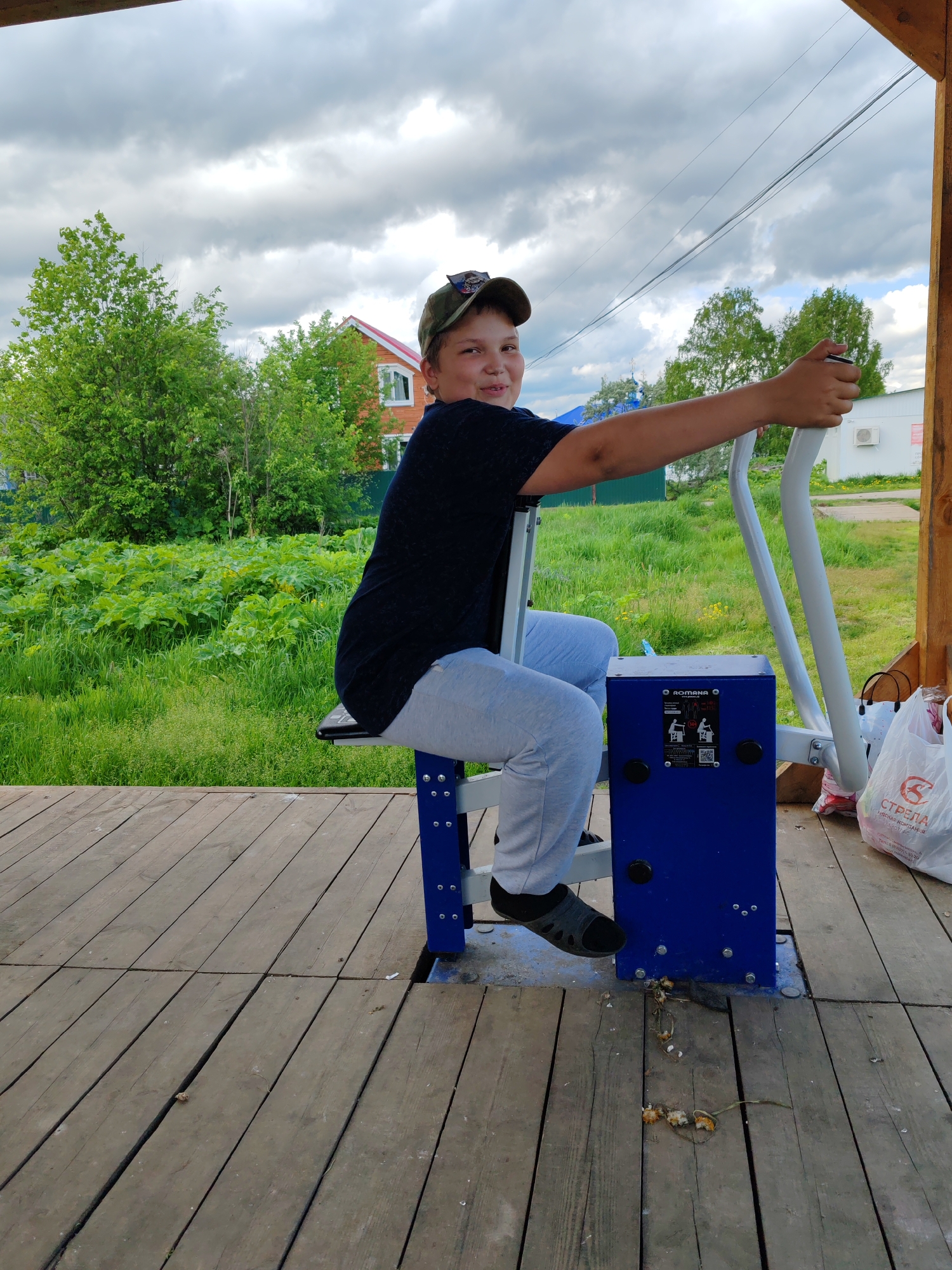 A person is seated on a blue and white outdoor exercise machine. The person is wearing a dark t-shirt, light-colored pants, and dark shoes. They also have a cap on their head. The exercise machine is placed on a wooden deck, and there appears to be debris on the floor under the machine. In the background, there is greenery including trees and a field, as well as buildings. The sky is cloudy. The location is Чухлэм, Russia, as stated. There is a bag visible to the right of the exercise machine with Cyrillic text on it.