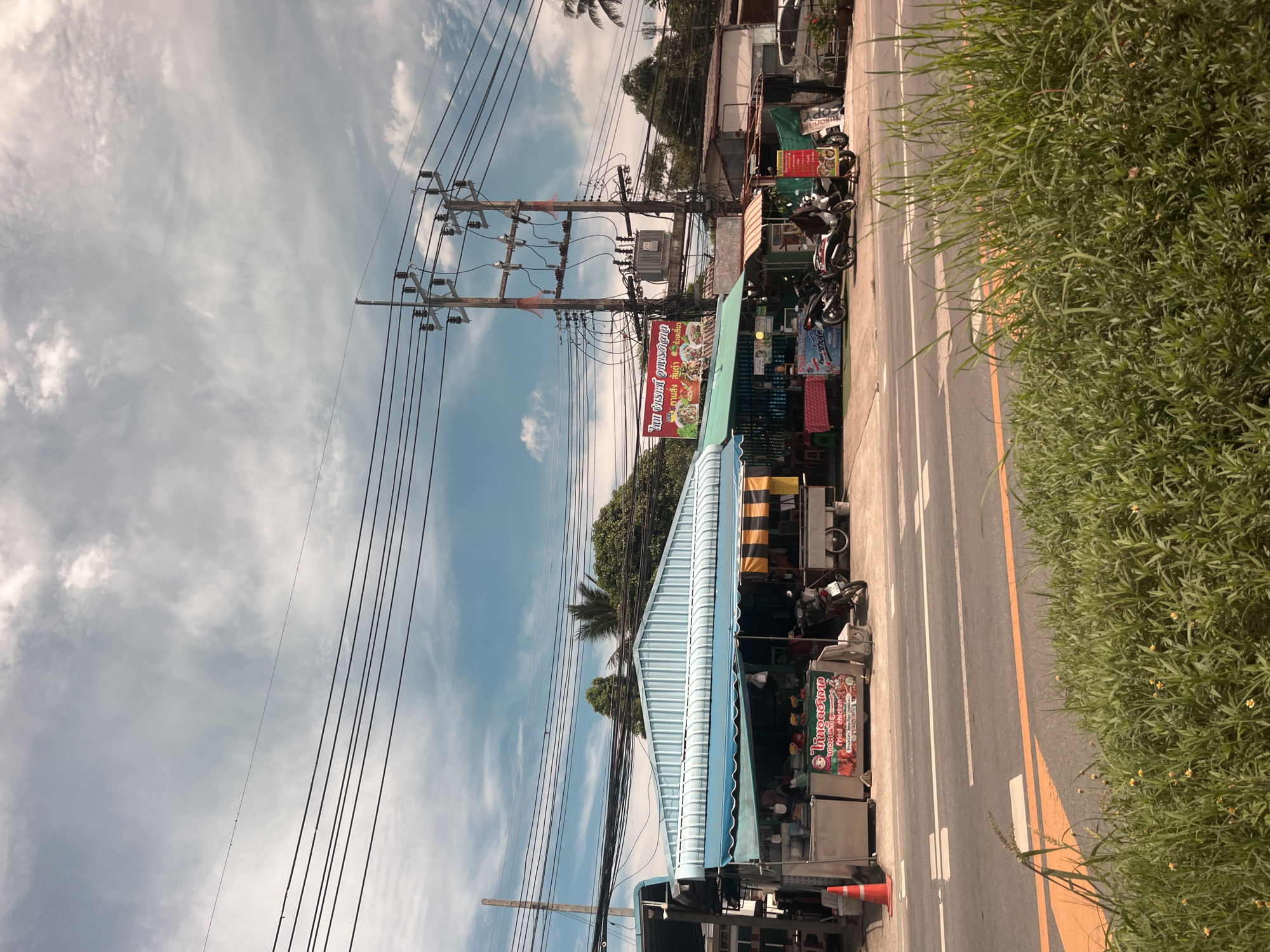 The image shows a street scene in Rawai, Thailand, with several food stalls set up along the roadside. The stalls have blue and green awnings and are selling various items, including what appears to be fried chicken. Several motorcycles are parked in front of the stalls. In the background, there are power lines and a transformer. The sky is cloudy. The road in the foreground is asphalt with yellow and white lane markings. Lush green grass and small yellow flowers are growing along the side of the road.