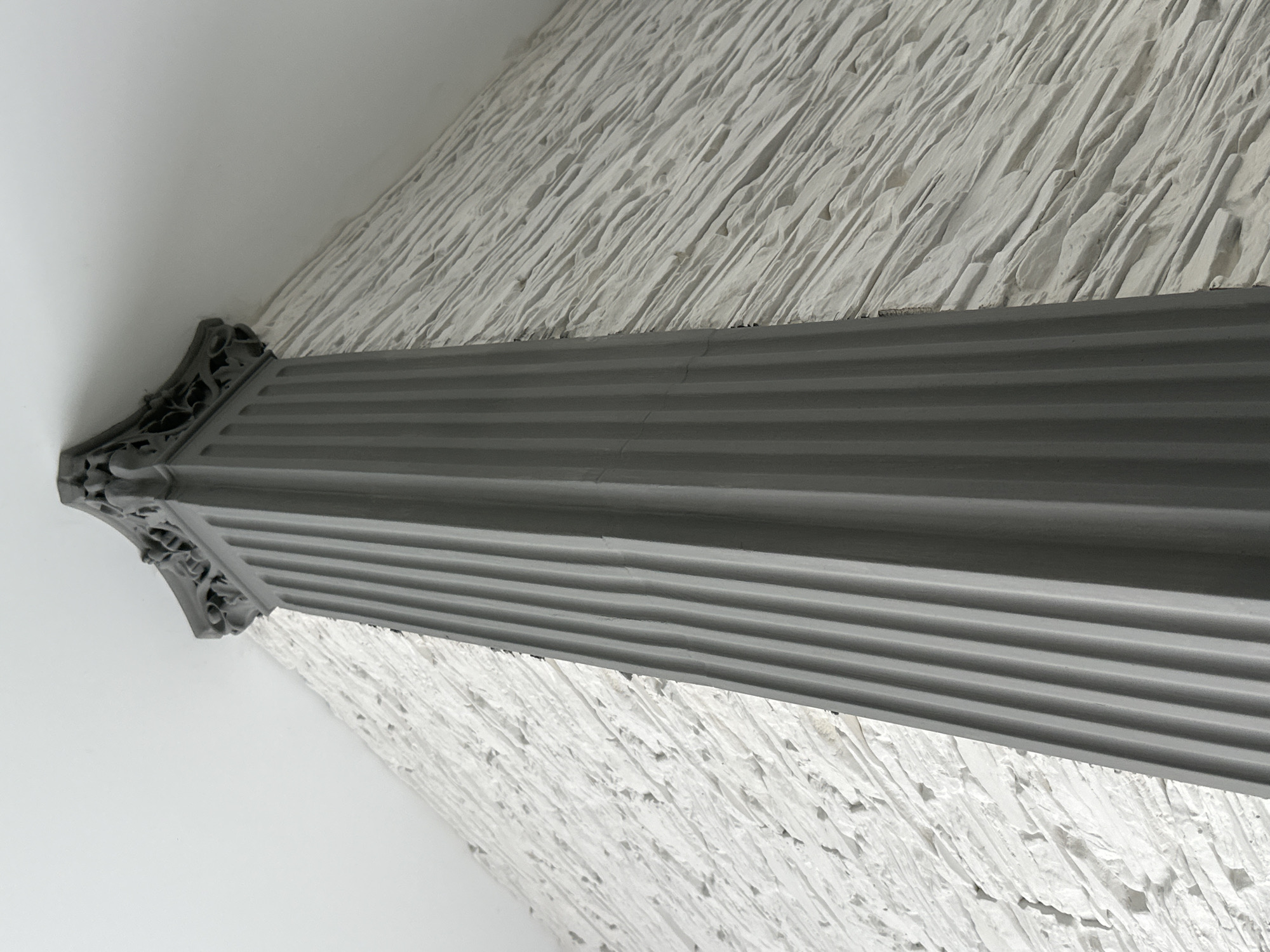 A low-angle shot depicts a gray, fluted column with an ornate capital, positioned against a textured, white wall. The background consists of the wall and a smooth, white surface adjacent to the column. The location is Unknown, Unknown.