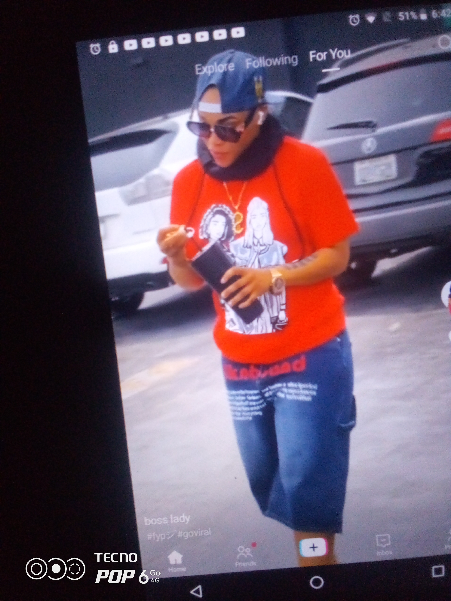 The image is a screenshot from a mobile device, displaying a social media feed. The feed shows a person wearing a red t-shirt, blue jeans, sunglasses, and a blue baseball cap. The person is holding a black object in their right hand. The background appears to be an outdoor urban environment, possibly a street or parking lot with vehicles visible. Text overlays on the screen include "Explore Following For You", "boss lady", "#fyp", "#goviral", "TECNO POP 6 4G", and navigation icons for "Friends", "Home", and "Inbox". The device displays a battery percentage of 51% and a time of 6:42. There is no information within the image to suggest the location is Howayi, Nigeria.
