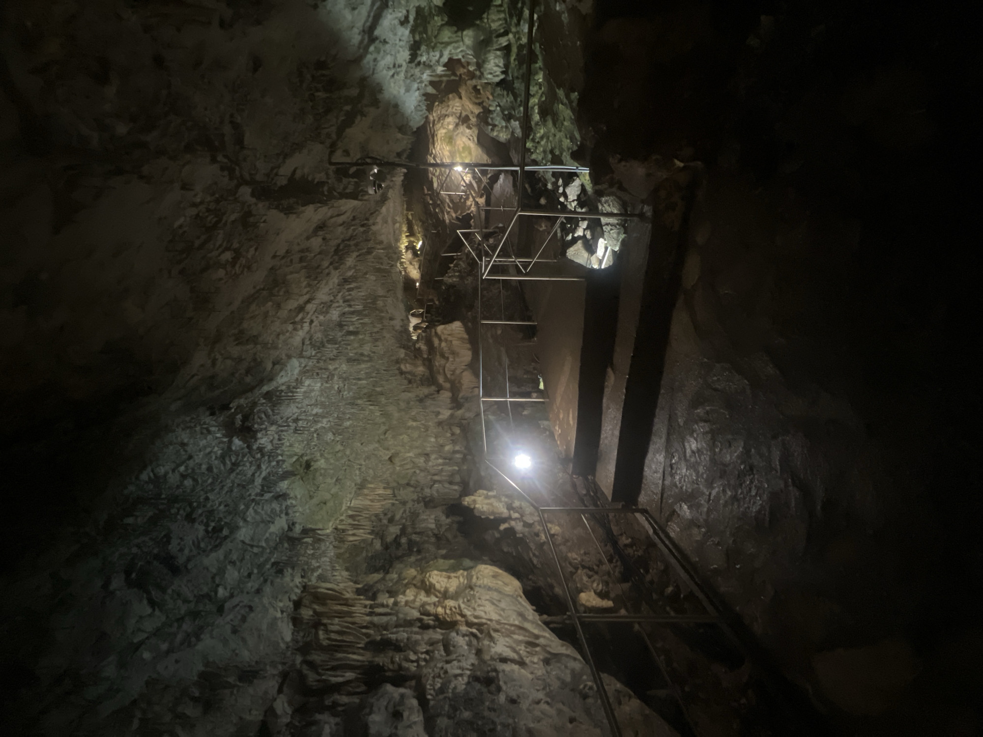 This image shows the interior of a cave system, featuring a metal walkway and stairs constructed to facilitate exploration. The cave walls are predominantly light gray and textured, with visible geological formations and mineral deposits. The lighting is dim, provided by artificial sources along the walkway, accentuating the uneven surfaces of the cave. The environment is dark and enclosed, typical of subterranean landscapes. The location is Unknown, Unknown.