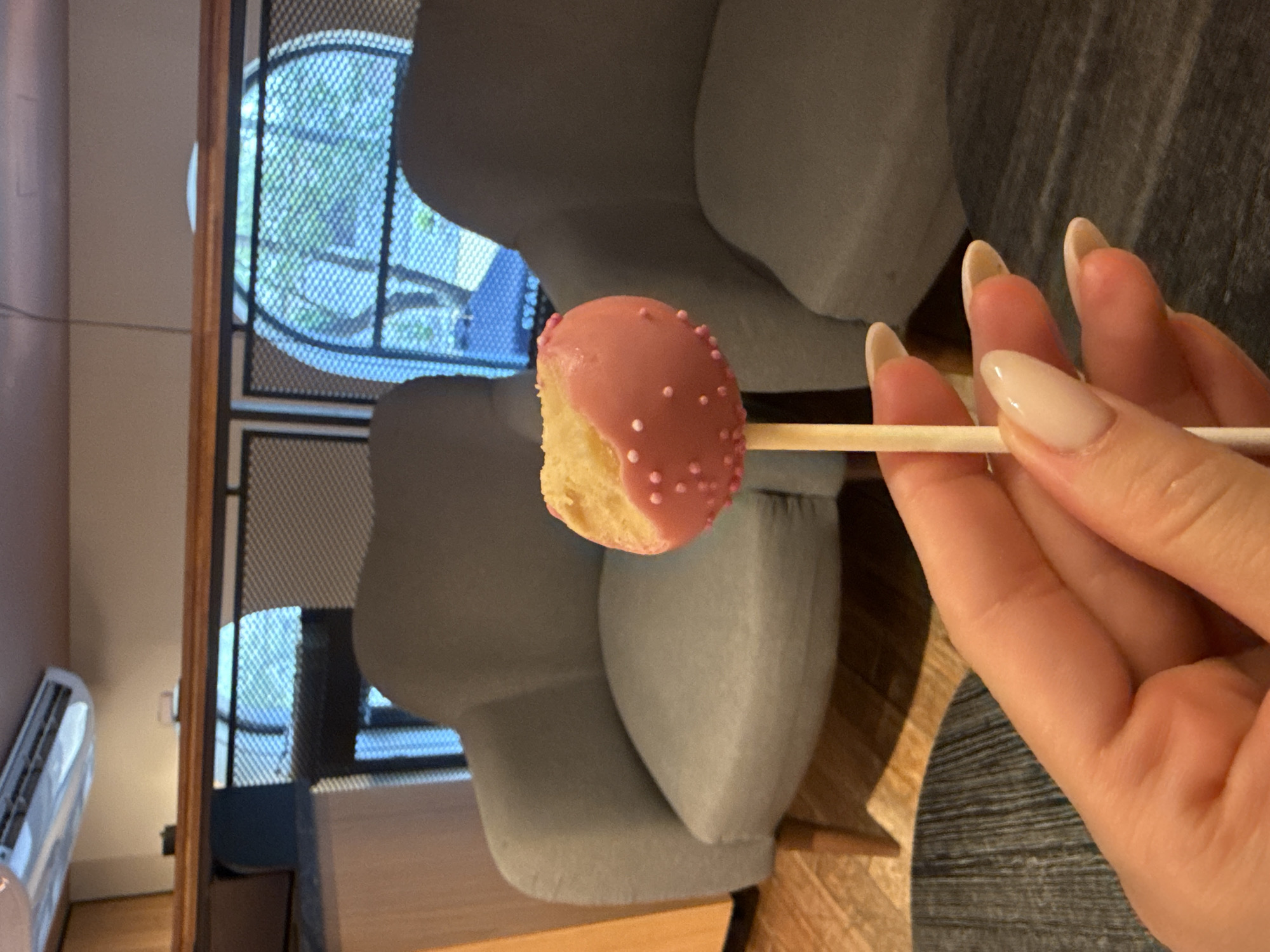 A close-up, portrait-oriented image, rotated 90 degrees clockwise, captures a person's hand holding a pink-frosted cake pop against an indoor background.
In the foreground, a right hand with long, light-colored, oval-shaped nails holds a wooden stick, from which a cake pop protrudes. The cake pop is half-covered in light pink frosting and adorned with small pink sprinkles. The visible cake portion beneath the frosting is a light yellow color. Below the hand, a warm-toned wooden surface, likely a table, is visible, and further down, a grey patterned rug or mat covers the floor.
The background features a modern indoor setting. To the left (which would be the top if the image were correctly oriented), a white air conditioning unit is mounted on a light-colored wall next to a brown door frame. Through a window with a mesh screen, lush green foliage from outside is visible, suggesting a daytime setting. In the middle ground, two large, grey upholstered chairs or lounge seats are prominent, their rounded forms contributing to the contemporary feel of the space. No text is visible in the image. The location context indicates this scene is in Sofia, Bulgaria.