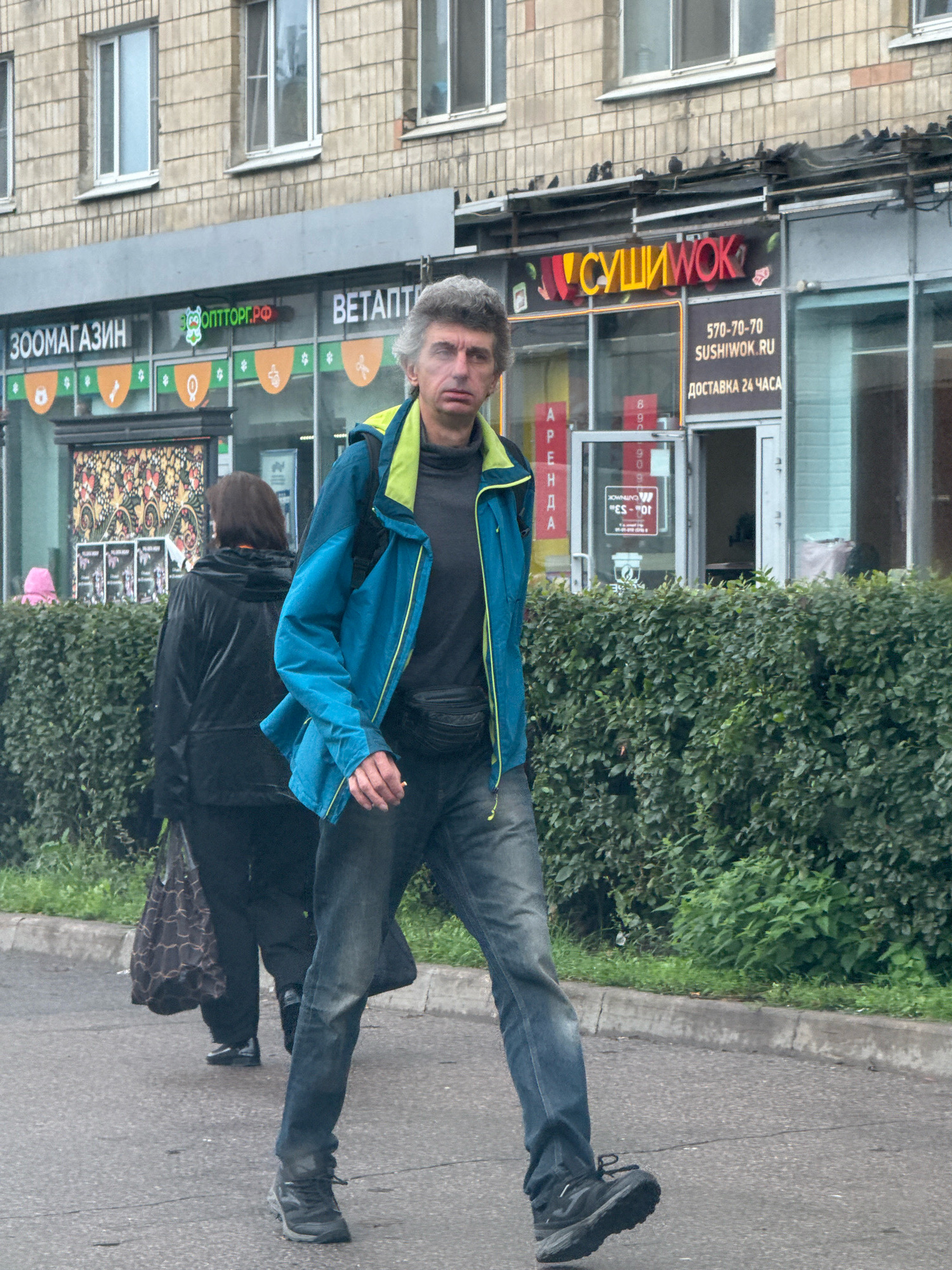 A medium-shot, eye-level, outdoor, daylight, full-body shot captures a Caucasian man walking on a paved street in front of a building with shops. The man, with greying hair and a serious expression, is wearing a blue and yellow jacket over a dark gray turtleneck, dark wash jeans, and black athletic shoes. He has a black fanny pack around his waist and is holding a lit cigarette in his right hand. He is walking from left to right, slightly angled towards the viewer. Behind him, a woman in a black jacket and black pants is walking away from the camera, carrying a patterned bag in her left hand. She is walking to the left, also on the paved street. The building behind them features several shop windows with signage, including "ЗОOMAGAZIN" and "СУШИWOK" in Cyrillic. The building's facade is made of beige brick with multiple windows. A dense green hedge runs along the sidewalk in front of the building. The overall environment appears to be a typical urban street scene in Saint Petersburg, Russia.
