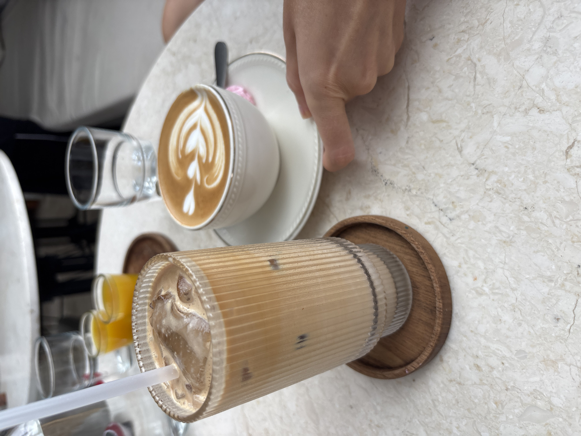 An eye-level, medium shot captures two drinks on a marble-topped table at a cafe, likely in Kuta Selatan, Indonesia. In the foreground, a tall glass with vertical ridges is filled with an iced coffee, complete with a white straw and ice cubes. This glass rests on a small, round wooden coaster. To the right, a white ceramic cup holds a latte with intricate latte art. The cup sits on a matching saucer, with a small pink item partially visible beneath it. A person's hand, with fingers extended and pointing, is visible on the right side of the frame, near the saucer. In the background, two small glasses of orange juice and an empty glass are also present on the table, along with glimpses of other cafe furniture.