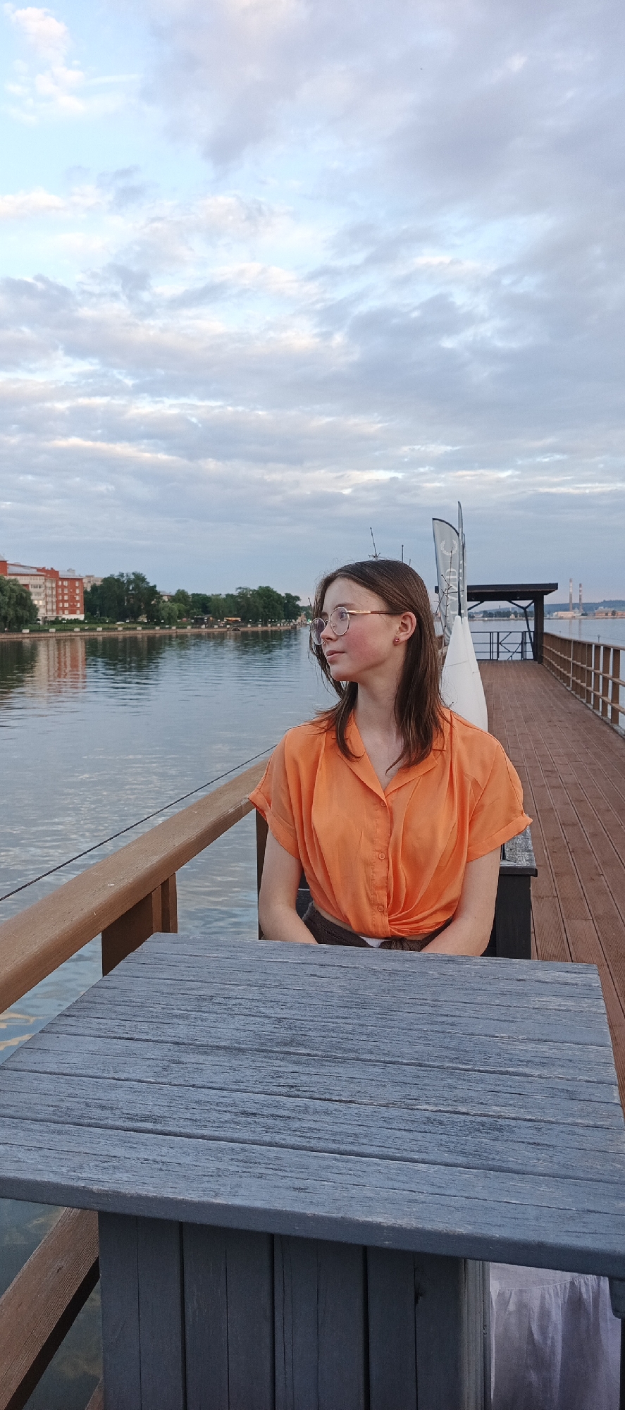 A young woman wearing glasses and an orange short-sleeved button-up shirt is seated at a wooden table on a pier. The table is a rustic blue color, and the pier is made of wooden planks. To her left, a brown wooden railing runs along the edge of the pier. Behind her, a calm body of water reflects the cloudy sky and a distant cityscape. The cityscape features buildings and trees on the opposite bank of the water. The sky is filled with a mix of light blue and scattered white and gray clouds. The scene appears to be in the city of Votkinsk, Russia, given the context of the request.