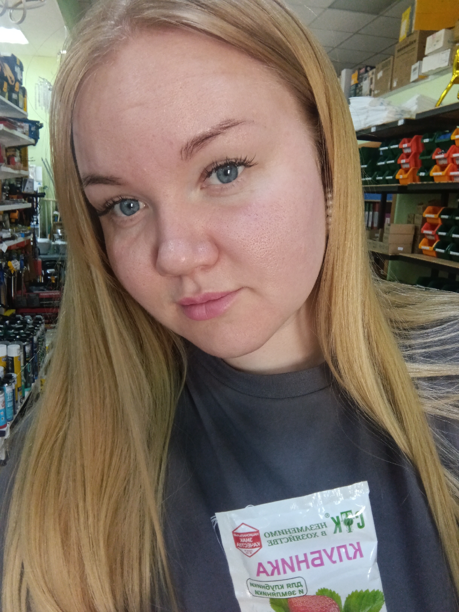A young woman with fair skin, light blue eyes, and long, straight, blonde hair is shown in a close-up selfie shot. She is wearing a gray t-shirt. The image is taken indoors, possibly in a shop, with shelves stocked with various tools, cans, and boxes visible in the background. She is holding a package of what appears to be strawberry-flavored snack or supplement, with text written in Cyrillic on it. The location, Фоки, Russia, is mentioned.