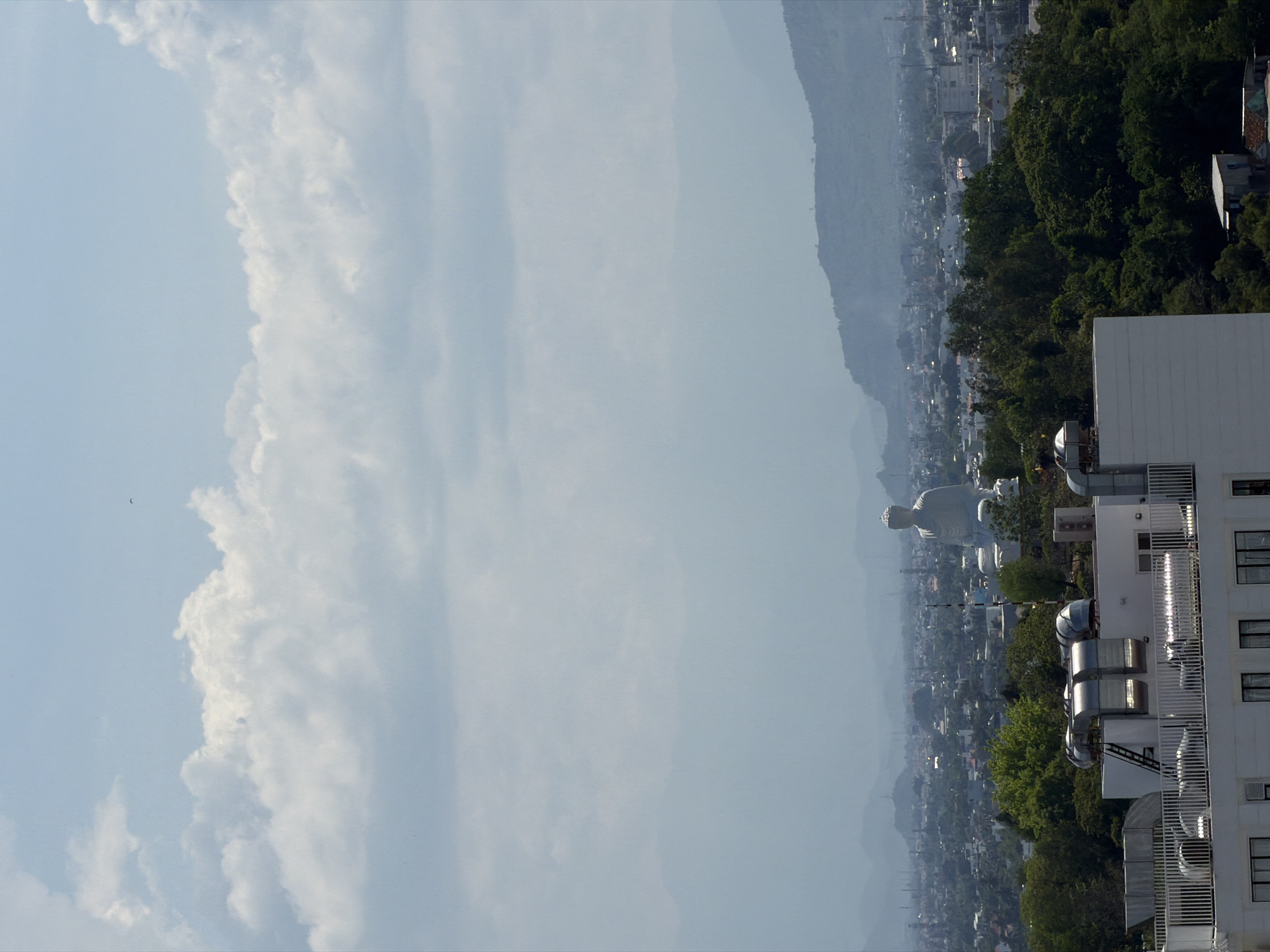This is an aerial photograph taken on a bright, overcast day, looking out over the city of Nha Trang, Vietnam. In the foreground, a modern, white building with a railing and air conditioning units on its roof is visible. Beyond this building, a dense, green forest separates the viewer from the city. In the midground, the white statue of a seated Buddha dominates the landscape, positioned on a hill overlooking the city. The city itself is a sprawling urban area with numerous buildings, some with red roofs, stretching towards the hazy mountains in the background. The sky is filled with large, white, cumulus clouds, and a faint haze or fog hangs over the distant mountains, obscuring details. There are no visible people or animals in the photograph, and no discernible actions or interactions are taking place. The overall impression is of a vast, panoramic view of a city and its surrounding natural landscape, with the prominent Buddha statue serving as a central focal point.