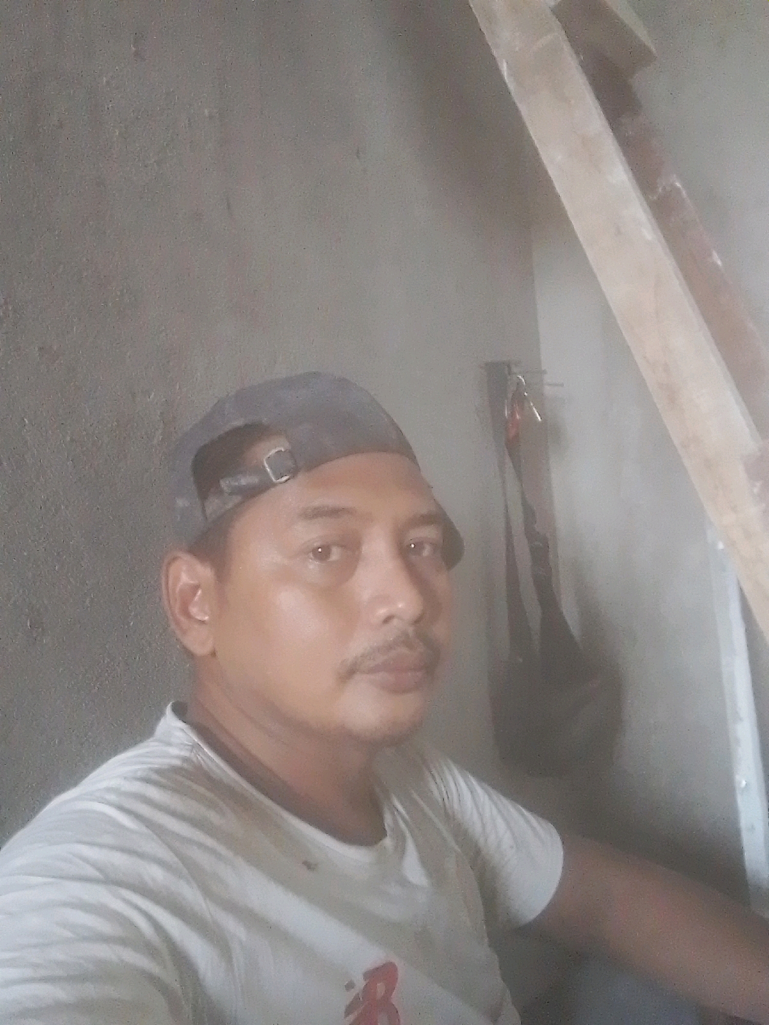 A medium shot depicts a man wearing a dark baseball cap and a white t-shirt. He is looking directly at the camera, with a mustache visible. The background consists of a textured gray wall on the left and a diagonal wooden structure on the right. A dark, bag-like object hangs on the wall to the right of the man. The lighting is somewhat diffused, with a slight haze. The overall setting appears to be an indoor or partially enclosed space, possibly a workshop or unfinished construction area. The location is West Jakarta, Indonesia.