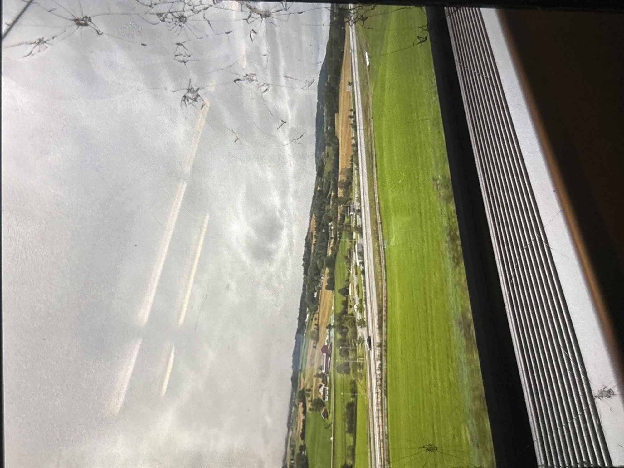 The image shows a view through a cracked window, likely from a train or bus, overlooking a rural landscape. The foreground features a gray, ribbed surface, possibly a window sill or a ventilation grate. Beyond this, a vibrant green field stretches out, transitioning into rolling hills with patches of yellow and green fields, interspersed with trees and a few scattered buildings with red roofs. A road, appearing to be a highway, runs horizontally across the middle ground, with a few vehicles visible. The sky is overcast with dramatic, cloudy formations. The window itself is covered in numerous cracks and some dirt or smudges, distorting the view. There is no indication of the city of Orihuela, Spain, or any urban elements.
