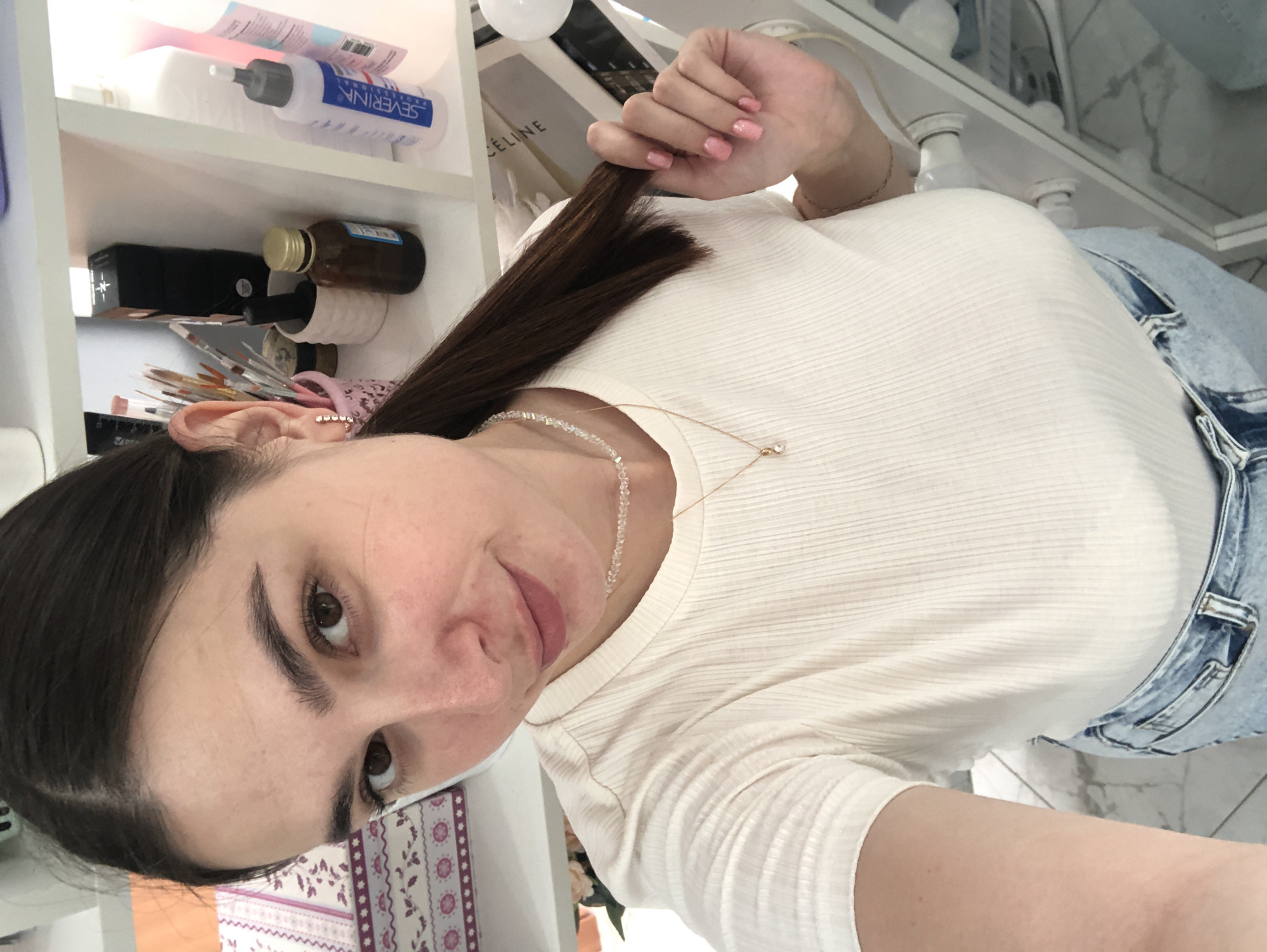 The image shows a young woman from the chest up, taking a selfie. She is wearing a cream-colored, ribbed t-shirt and blue jeans. Her long, dark brown hair is pulled to the side and she is holding a strand of it with her right hand. Her nails are painted a light pink. She has on a delicate necklace with clear beads and a small pendant. The background appears to be a shelf unit filled with various products, possibly in a bathroom or vanity area. Some items visible on the shelves include bottles of lotions or toners, a black box, and makeup brushes. The overall lighting suggests it could be daytime, possibly indoors with artificial light. The woman's expression is neutral, and the image is taken at a slightly upward angle, typical of a selfie. There is text visible on a white bottle in the background that reads "SEVERINA PROFESSIONAL".