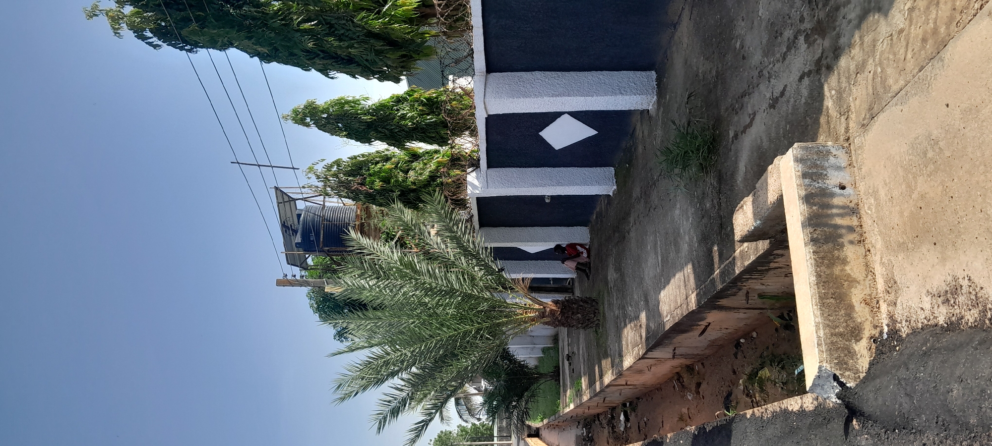 In Jalingo, Nigeria, a paved walkway runs alongside a drainage ditch. A person is seated against a dark blue and white patterned wall topped with barbed wire. A palm tree stands in front of the wall. In the background, a large black water tank is visible on a raised platform, and power lines cross the sky. The scene is bathed in natural daylight.