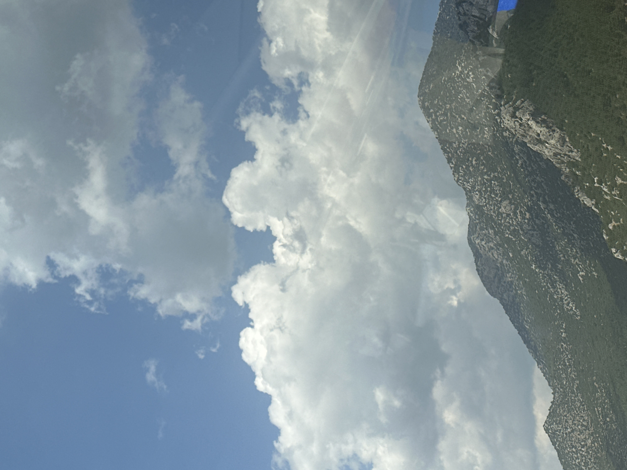 The image shows a view of a mountainous landscape under a sky filled with fluffy white and grey clouds. The mountains are covered in green vegetation and rocky outcrops, with a prominent peak in the foreground. The sky is a bright blue, with sunlight streaming through some of the clouds, creating a lens flare effect. A small blue rectangular object is visible on the far right side of the image, partially obscuring the mountain. The image appears to be a photograph taken from a distance, possibly from a vehicle or a higher vantage point. The location is identified as Korkuteli, Turkey.