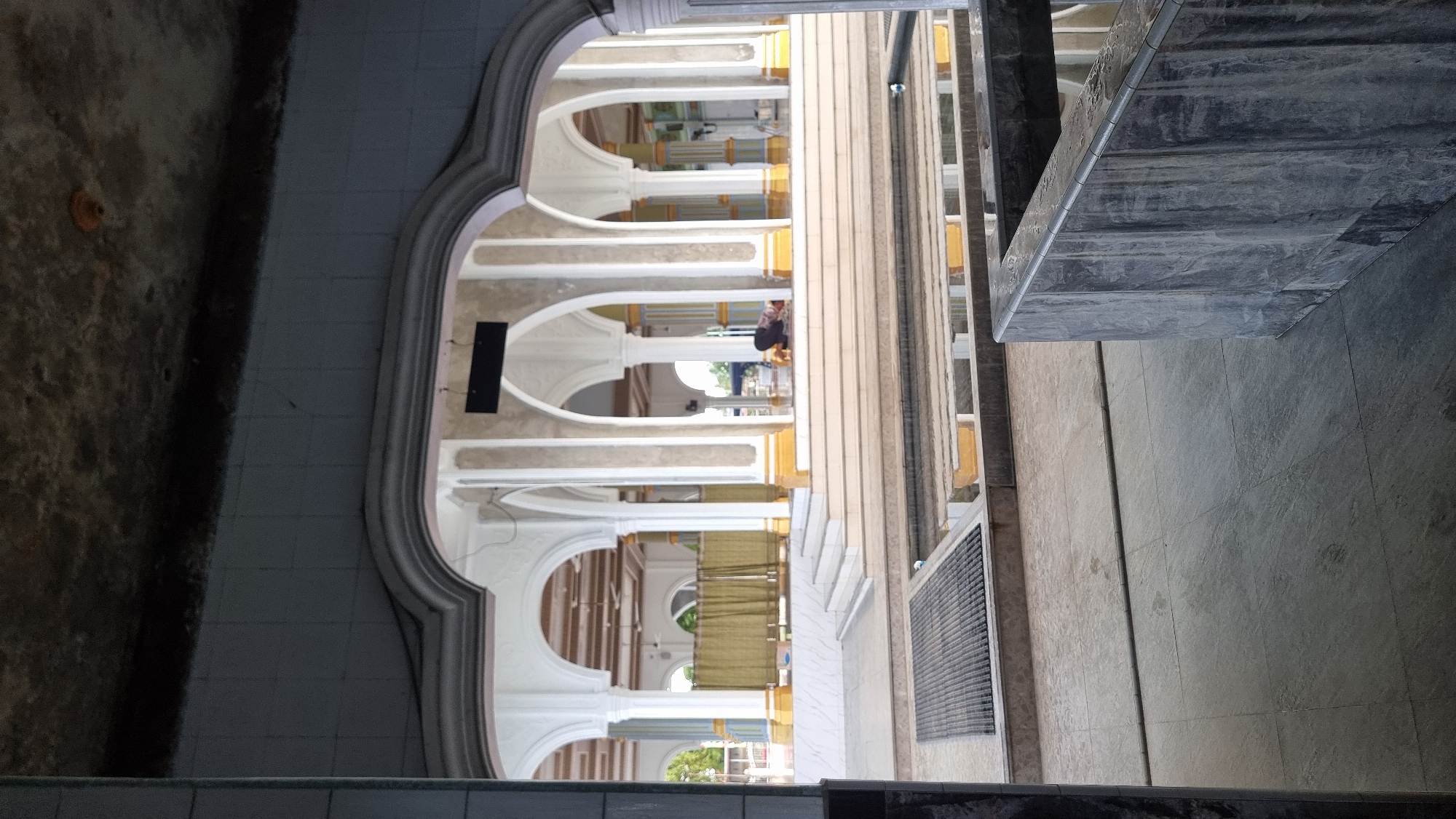 This image depicts the interior of a mosque, likely in Meunasahlhok, Indonesia, as indicated by the architectural style. The scene is viewed from an entrance archway looking into the main prayer hall. Multiple arched colonnades, supported by grey and white pillars with yellow accents, extend into the background. A person is visible in the distance, kneeling on the floor in prayer. Green curtains hang behind a section of the colonnade on the left. In the foreground, a water feature or ablution area with a grate is present. The overall environment suggests a place of worship with traditional Islamic architectural elements.
