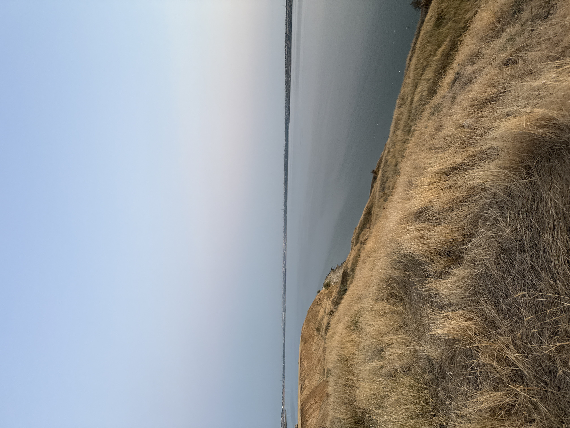 A wide, calm body of water stretches across the mid-ground and background of the image, meeting a distant, hazy shoreline with faint structures. The water reflects the pale, clear sky above, which graduates from a soft blue at the top to a lighter, almost white hue near the horizon. In the foreground and descending into the left side of the frame is a steep, dry, grassy slope. The grass is a golden-brown color, indicative of dry vegetation. The terrain appears rugged and natural, with evidence of erosion visible on the slope leading down towards the water. The overall scene suggests a natural landscape with a distant urban or industrial presence along the far shore.