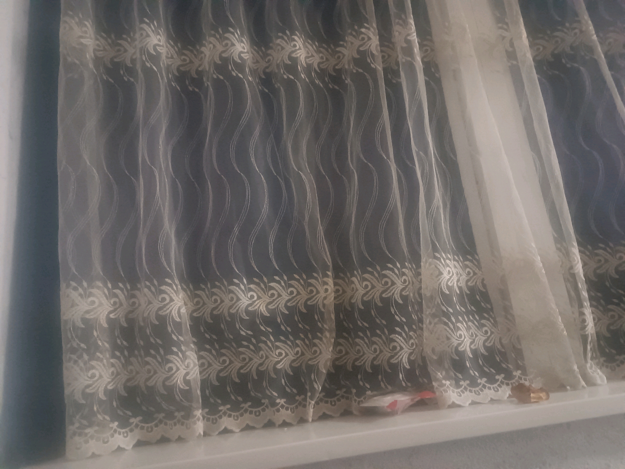 The image shows a close-up view of a windowsill with curtains. The curtains are made of sheer white fabric with an embroidered floral pattern along the bottom edge and vertical wavy lines. A portion of a blue curtain is visible behind the sheer curtain. There are some small, indistinct objects on the windowsill, appearing to be plastic wrappers or similar debris. The scene is indoors, likely a room with a window. The location is specified as Кенжекул, Kyrgyzstan.