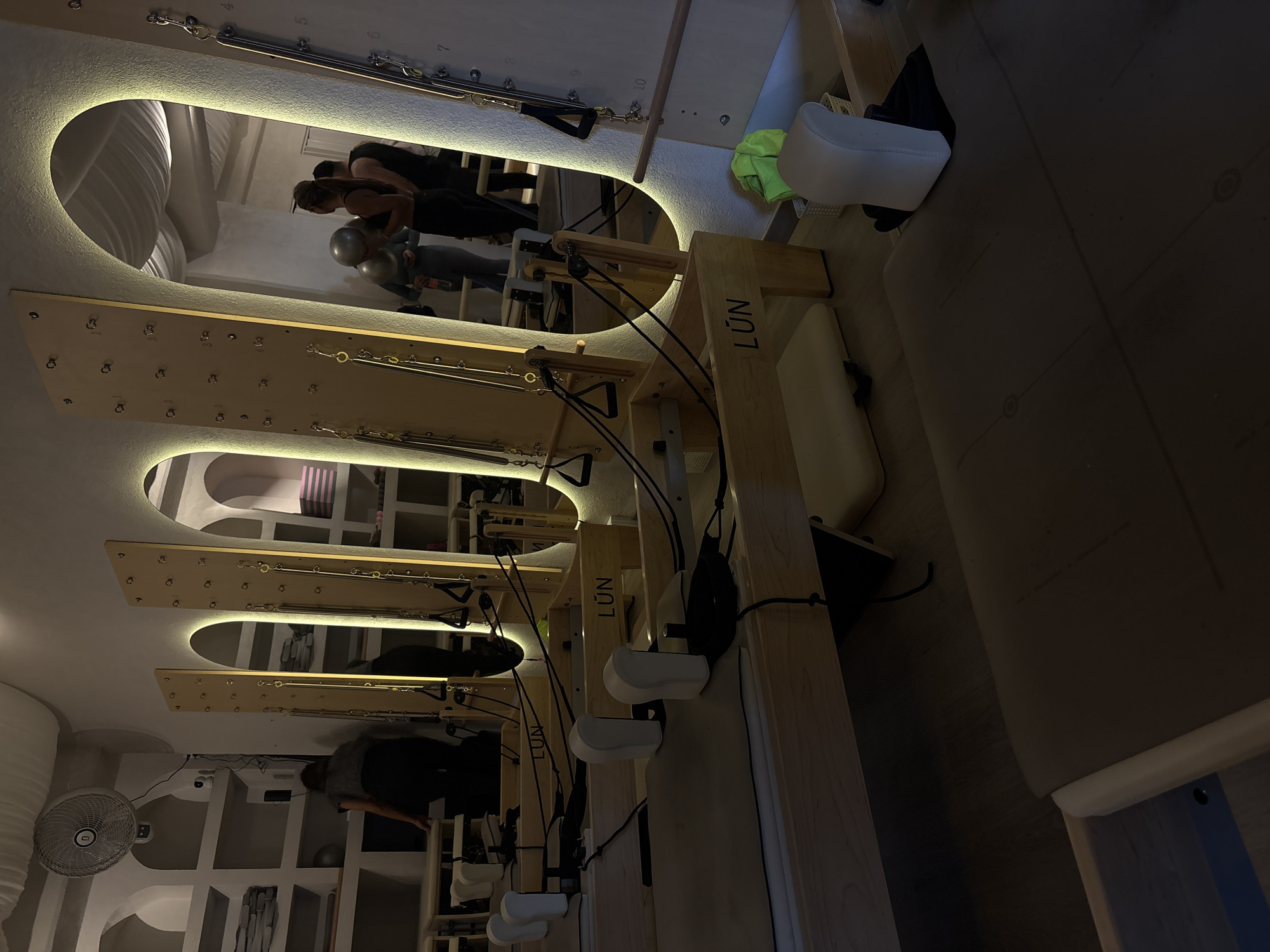 The image shows a Pilates studio in Mexico City, Mexico. There are four Pilates reformer machines, positioned parallel to each other. Each machine consists of a sliding carriage on a track, with a foot bar, shoulder rests, and straps. The studio has a minimalist design with white walls and recessed lighting that illuminates the reformers from underneath. The walls appear to be made of concrete. There is a fan on the wall. People are exercising on the machines.