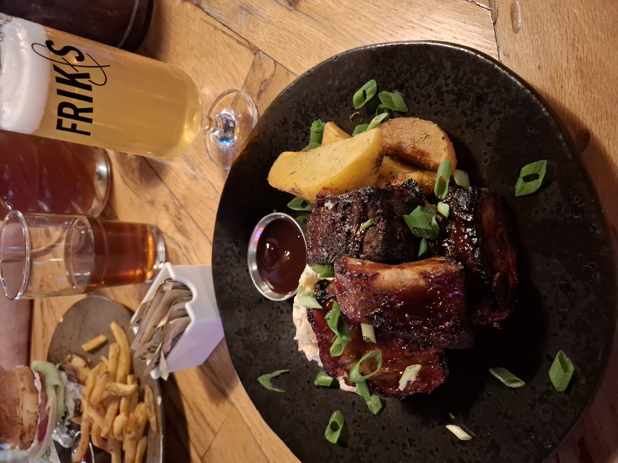 A close-up shot shows a black plate of ribs and potatoes. The ribs are glazed with a dark sauce and garnished with chopped green onions. The potatoes are wedge-shaped and appear roasted. A small bowl of dark sauce is also on the plate. In the background, a glass of beer with the logo "FRIKS" is visible, along with another glass of amber liquid and a burger with fries on a separate plate. The setting appears to be a restaurant with a wooden table.