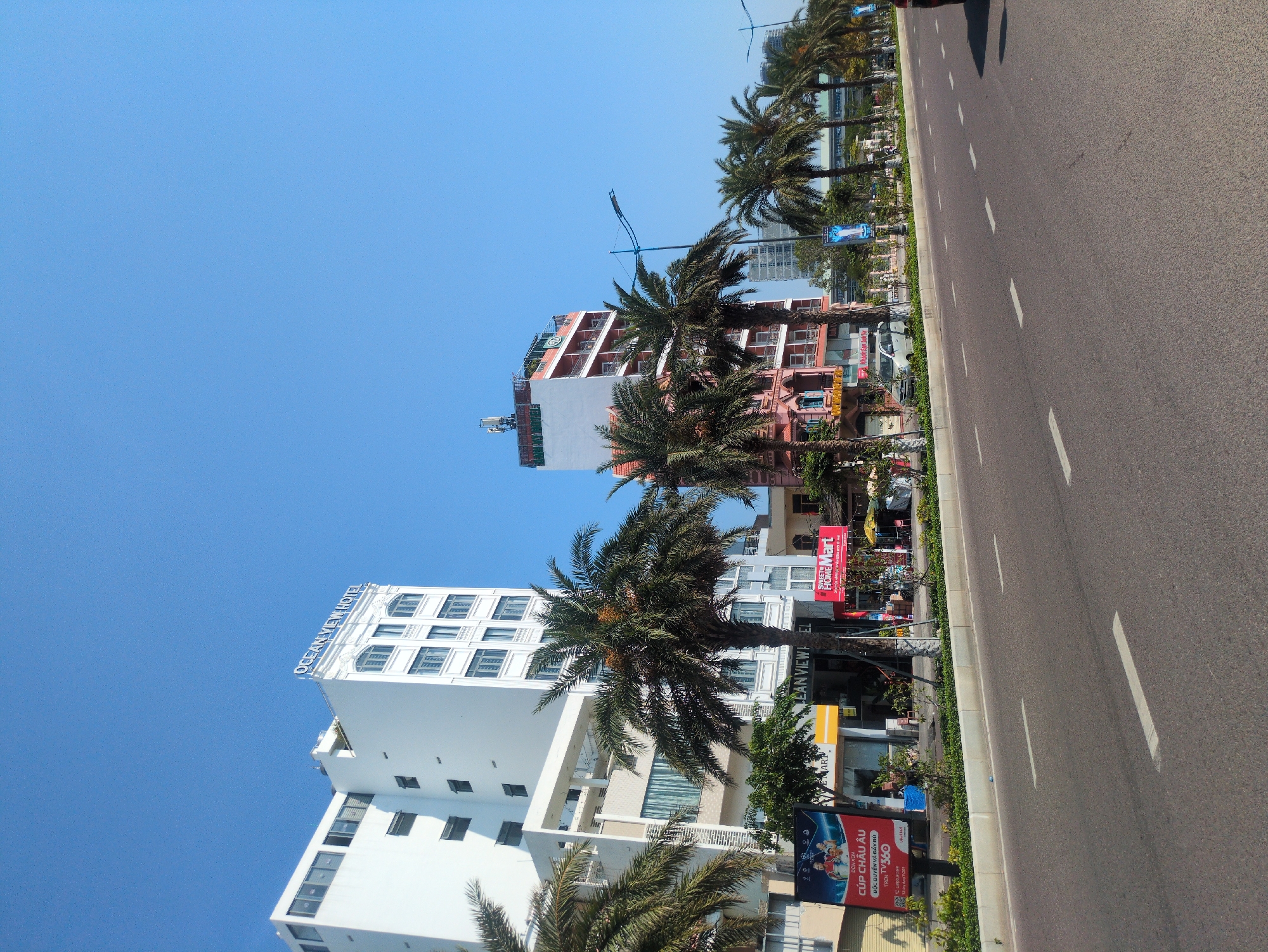 The image captures a sunny urban scene in South Quy Nhon Ward, Vietnam. A wide asphalt road dominates the foreground, marked with solid white lane dividers. A sidewalk borders the road, lined with palm trees and various signage. Buildings stand along the sidewalk, including the prominent white "Ocean View Hotel," a multi-story structure. There is a red “HOME Mart” sign and several advertisements, including one for a European Cup. The clear blue sky fills the upper portion of the frame.